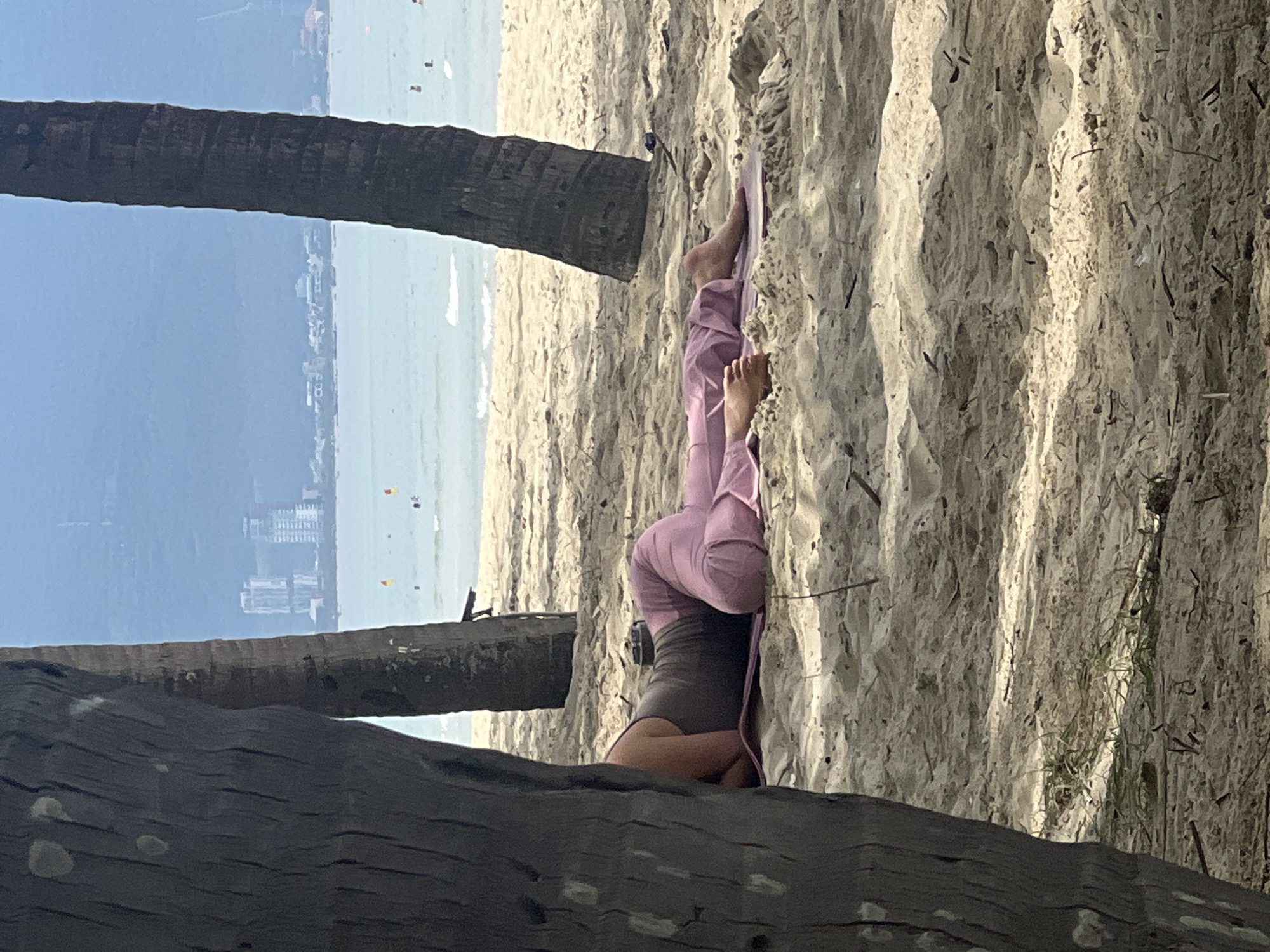 The image depicts a serene beach scene in Da Nang, Vietnam, likely My Khe Beach, known for its expansive sandy shores and calm waters. In the foreground, the rough textures of palm tree trunks partially obscure the view, framing the central subject. A person is lying on a yoga mat on the sandy beach, facing away from the camera and towards the ocean. They are wearing a grey tank top and loose-fitting pink pants, with their legs bent. Their feet are visible, resting on the mat.
The middle ground reveals the gentle waves of the sea meeting the shore, with several small figures of people swimming or wading in the water. In the distance, a cityscape is visible, characterized by a cluster of high-rise buildings that suggest the urban proximity of Da Nang. The hazy atmosphere softens the details of the city and the distant mountains, creating a tranquil and slightly dreamlike quality. The lighting suggests it is daytime, with diffused sunlight casting soft shadows on the sand. The overall impression is one of peaceful relaxation amidst a beautiful coastal environment.