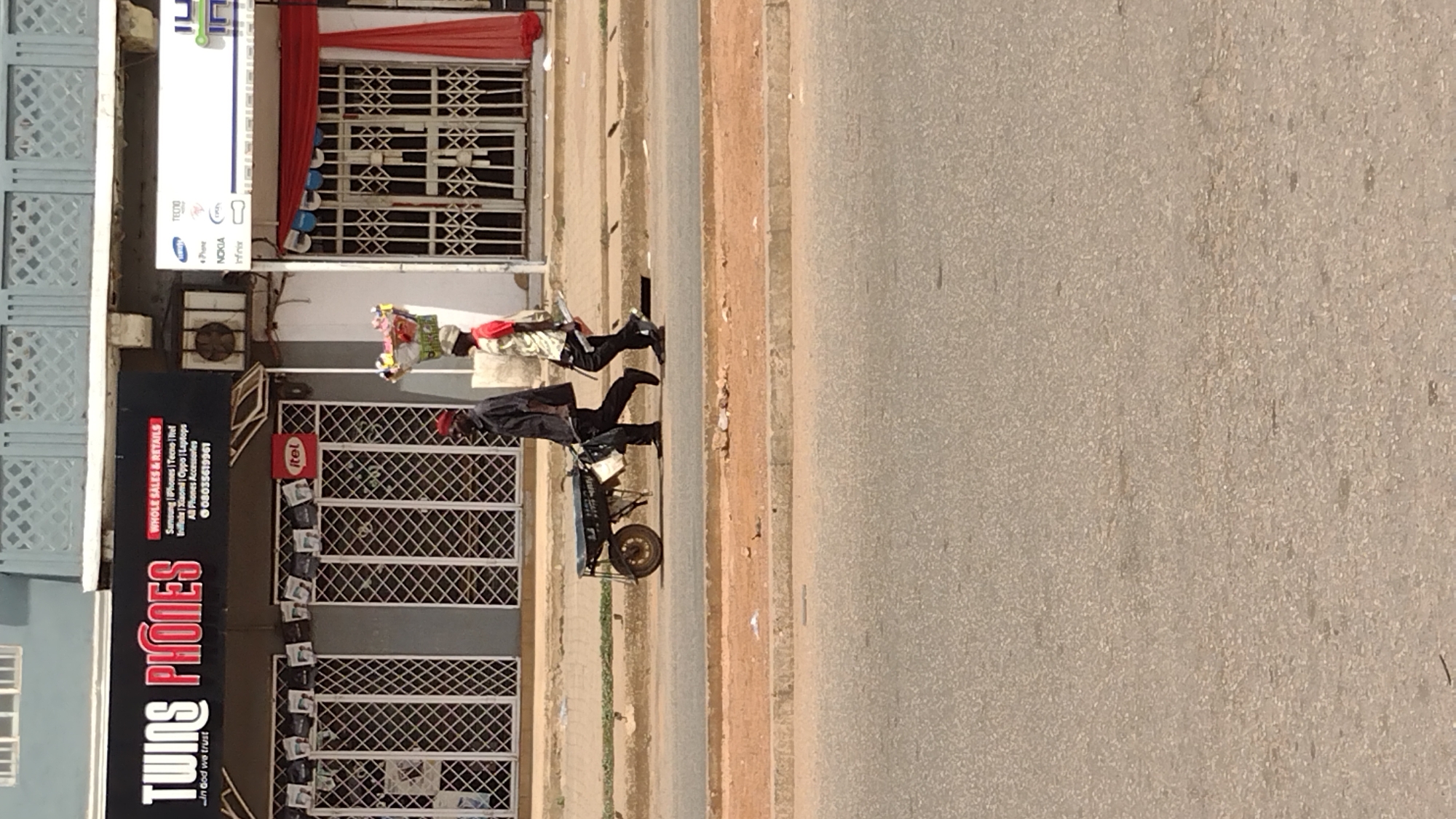 The image is a daytime outdoor scene apparently showing a street in Jos, Nigeria. On the left side of the frame is the storefront of a shop called "TWINS PHONES," with a sign advertising the sale of various phone brands including Samsung, Tecno, Itel, Infinix, and Oppo. A small sign indicates that they sell laptops and phone accessories. A gate covers the storefront. Two people are walking along the sidewalk. The first is pulling a small wheeled cart. The second person is carrying what appears to be a large bundle of goods on their head. The street fills the right side of the frame and appears to be mostly empty.
