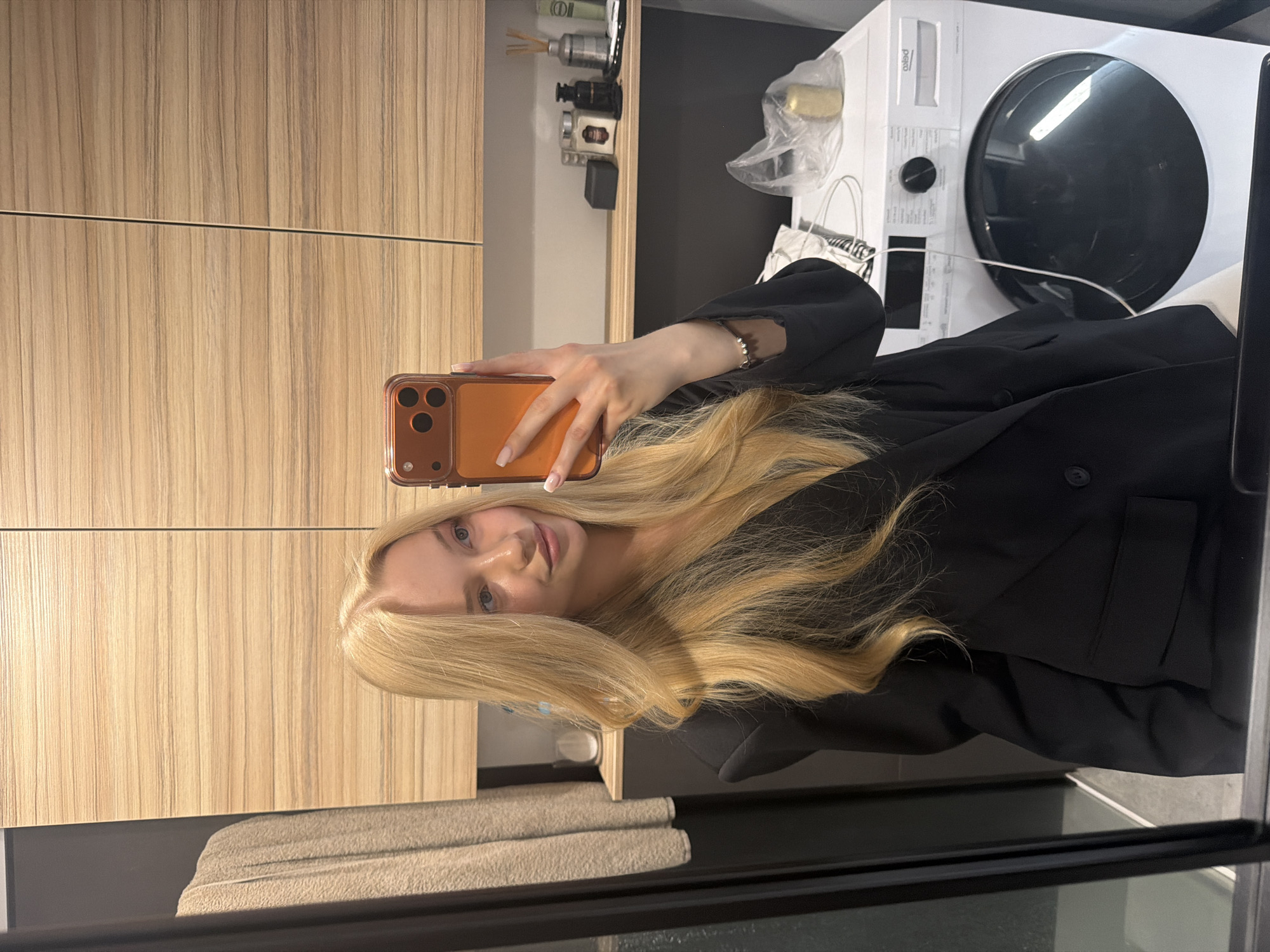 A woman is taking a selfie in what appears to be a bathroom. The location is Unknown, Unknown. She is holding an orange smartphone in her right hand, visible in the upper-left corner. The woman has long blonde hair and is wearing a black blazer. To the left of her is a light brown wooden cabinet, and to the right is a white appliance, possibly a washing machine or dryer. There are miscellaneous items on top of the cabinet, including what appears to be a decorative scent diffuser and other containers. The photo appears to be taken in front of a mirror.