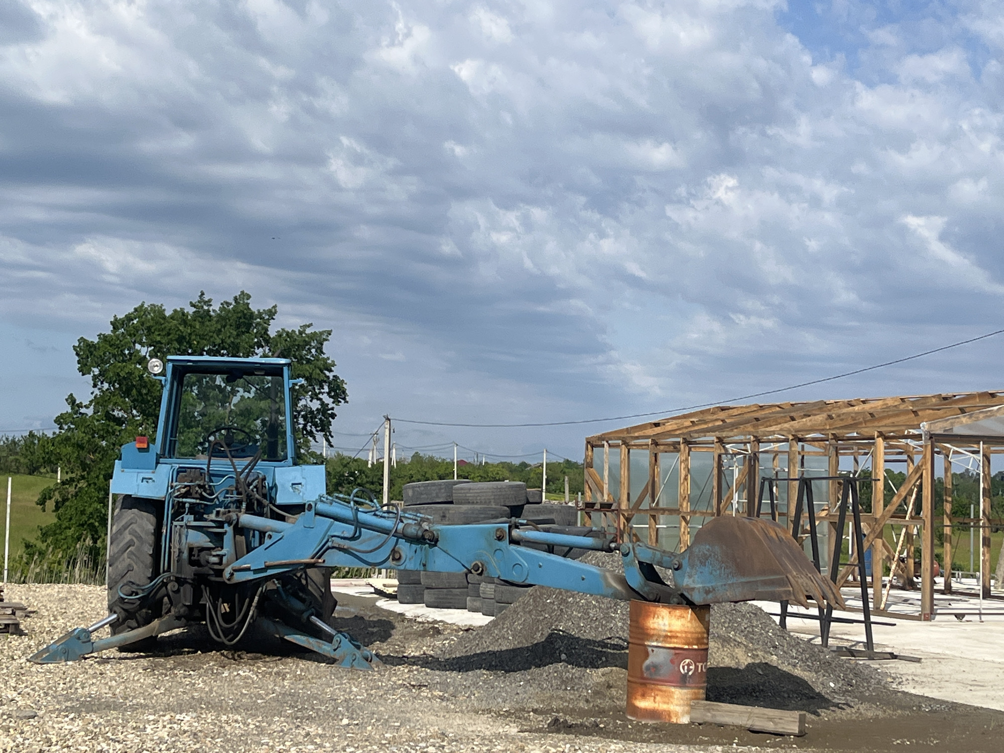 The image depicts a blue tractor with a front-end loader attachment parked in an outdoor area. Behind the tractor, there is a stack of used tires and a pile of gravel. An orange, rusted barrel with a logo is located near the loader bucket. In the background, there is a partially constructed wooden frame structure, possibly a greenhouse or shed. The ground is covered with gravel and stones. A tree is visible to the left of the tractor. The sky is cloudy. The location is Chetuk, Russia.