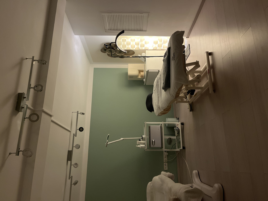 A massage room features a treatment table with a blanket, adjustable headrest, and side supports. Medical equipment includes a sterilizer cabinet with shelves, an adjustable lamp, and a footrest wrapped in a towel. The room's décor is minimalist, with light green walls and a white ceiling. Decorative lighting fixtures adorn the walls. A vent is visible on the ceiling. The floor is light brown.
