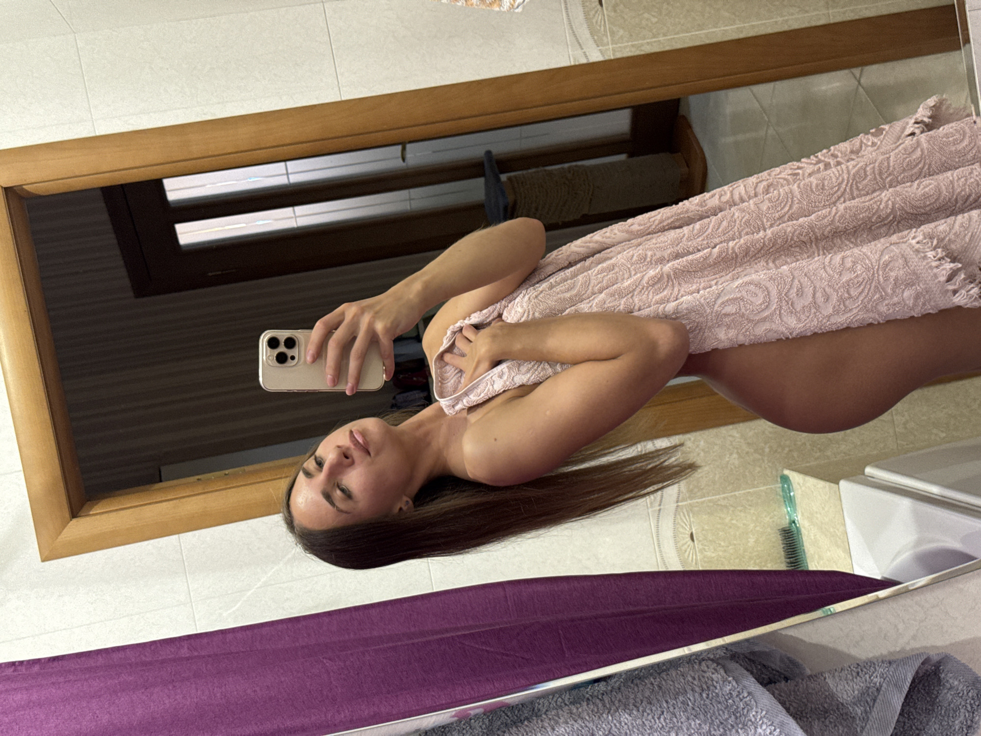 A young woman with long brown hair is taking a mirror selfie in a bathroom. She is holding a pink towel, which covers her body. She is wearing a ring on her left ring finger. The bathroom appears to be in an apartment in Saint Petersburg, Russia.