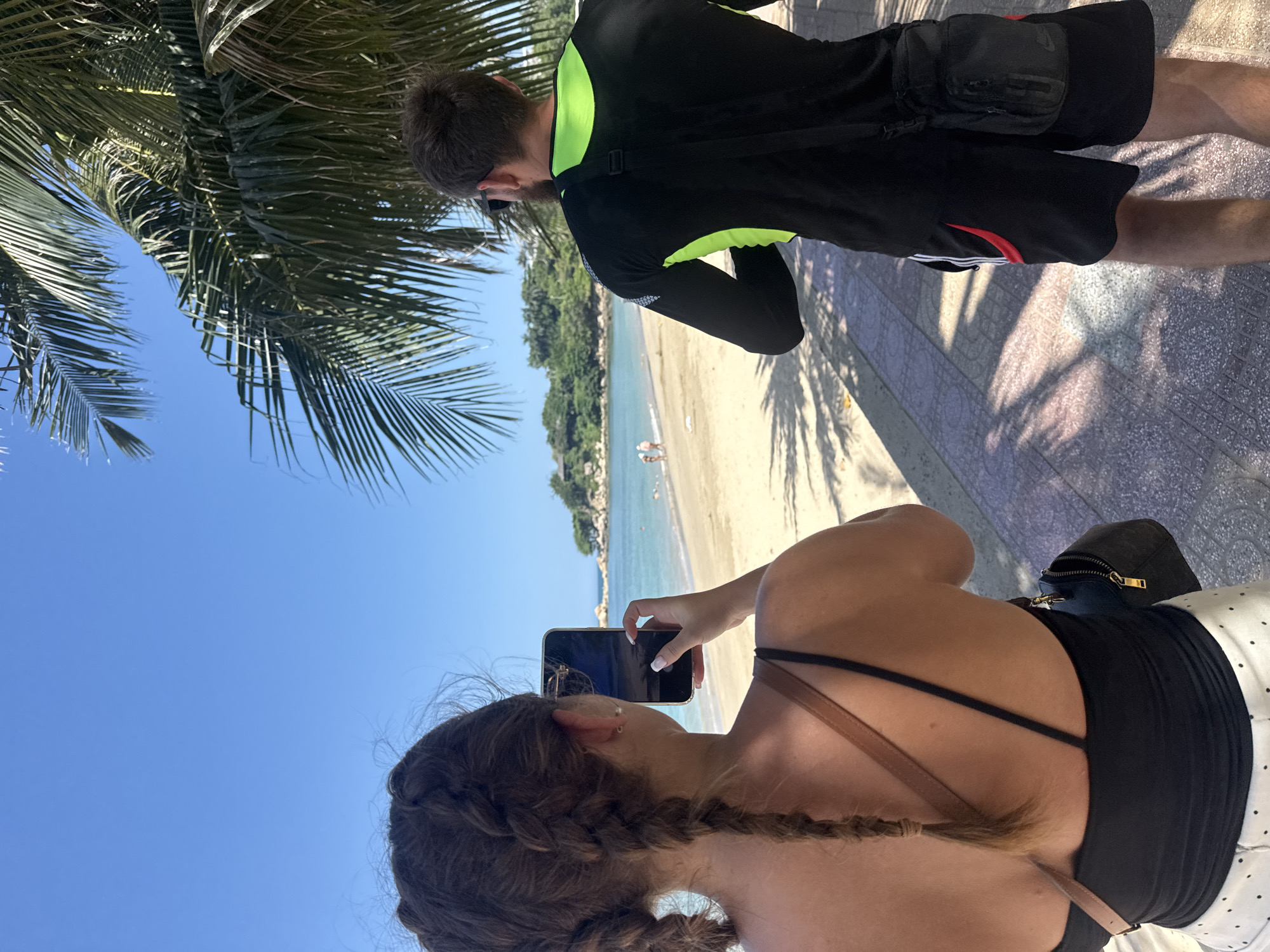 In Nha Trang, Vietnam, two individuals are depicted on a beach promenade under a clear blue sky. The woman, seen from behind, has her hair styled in two braids and is holding a smartphone, seemingly taking a photo of the beach and the ocean. She is wearing a black tank top and white polka-dot pants. A small black purse is slung across her shoulder. To her right, a man with a beard and sunglasses is also looking towards the sea. He is wearing a black long-sleeved rash guard with neon green accents and black athletic shorts. He has a small black bag with a strap across his chest. The beach itself is sandy, with calm turquoise water and a few distant figures wading near the shore. Lush green vegetation and trees, including palm trees casting shadows on the promenade, frame the scene.
