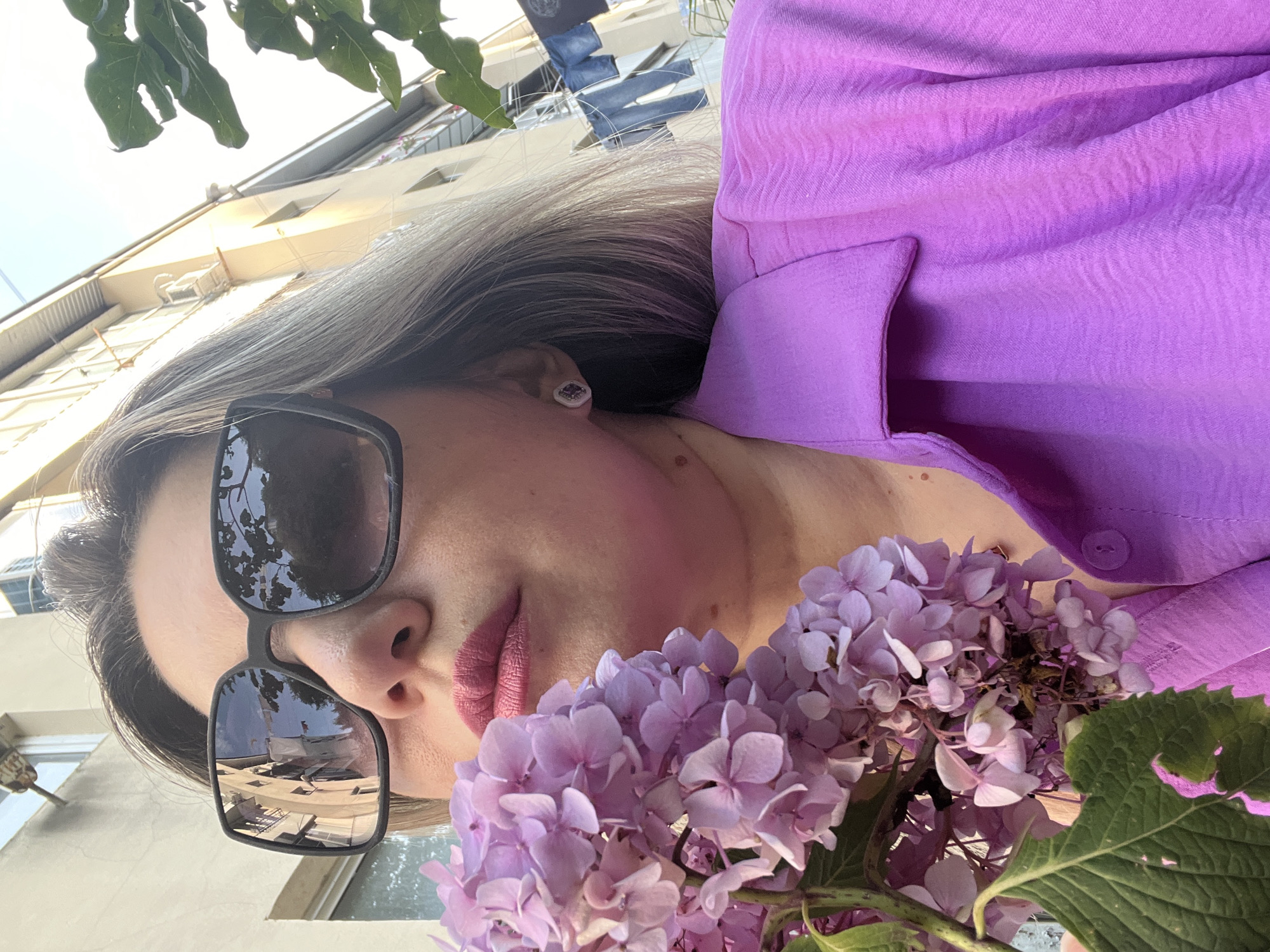 The image is a close-up, outdoor selfie of a woman with blonde hair, wearing sunglasses and a vibrant purple shirt. She is holding a cluster of light purple hydrangeas near her face. The woman is wearing subtle makeup, with pink lipstick and blush. A small stud earring is visible on her left ear. The sunglasses reflect a building with multiple windows and what appears to be clothes hanging on a line outside, suggesting a residential or urban environment. In the background, out-of-focus green leaves from a tree hang from the top of the frame, and a light-colored building with windows is visible. The overall lighting suggests it is daytime, likely sunny, casting shadows and highlighting the textures of the shirt and flowers. The woman's pose and the flowers create a relaxed and pleasant mood. The location context is Sochi, Russia.
