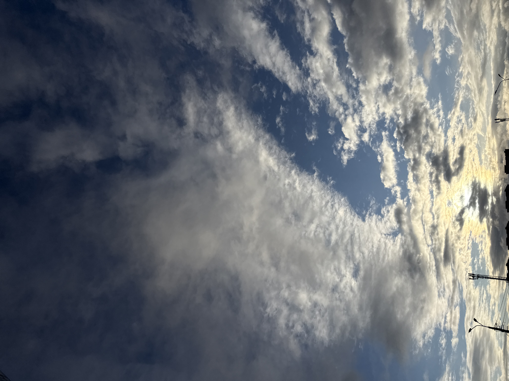 The image captures a dynamic sky in Krasnoyarsk, Russia. The sky transitions from a deep blue on the left to a lighter blue interspersed with elongated, streaky clouds on the right. These clouds are predominantly white and gray, some catching the light and appearing brighter. On the right edge, the silhouettes of buildings, streetlights, and a telecommunications tower are visible against the bright sky, indicating an urban setting.