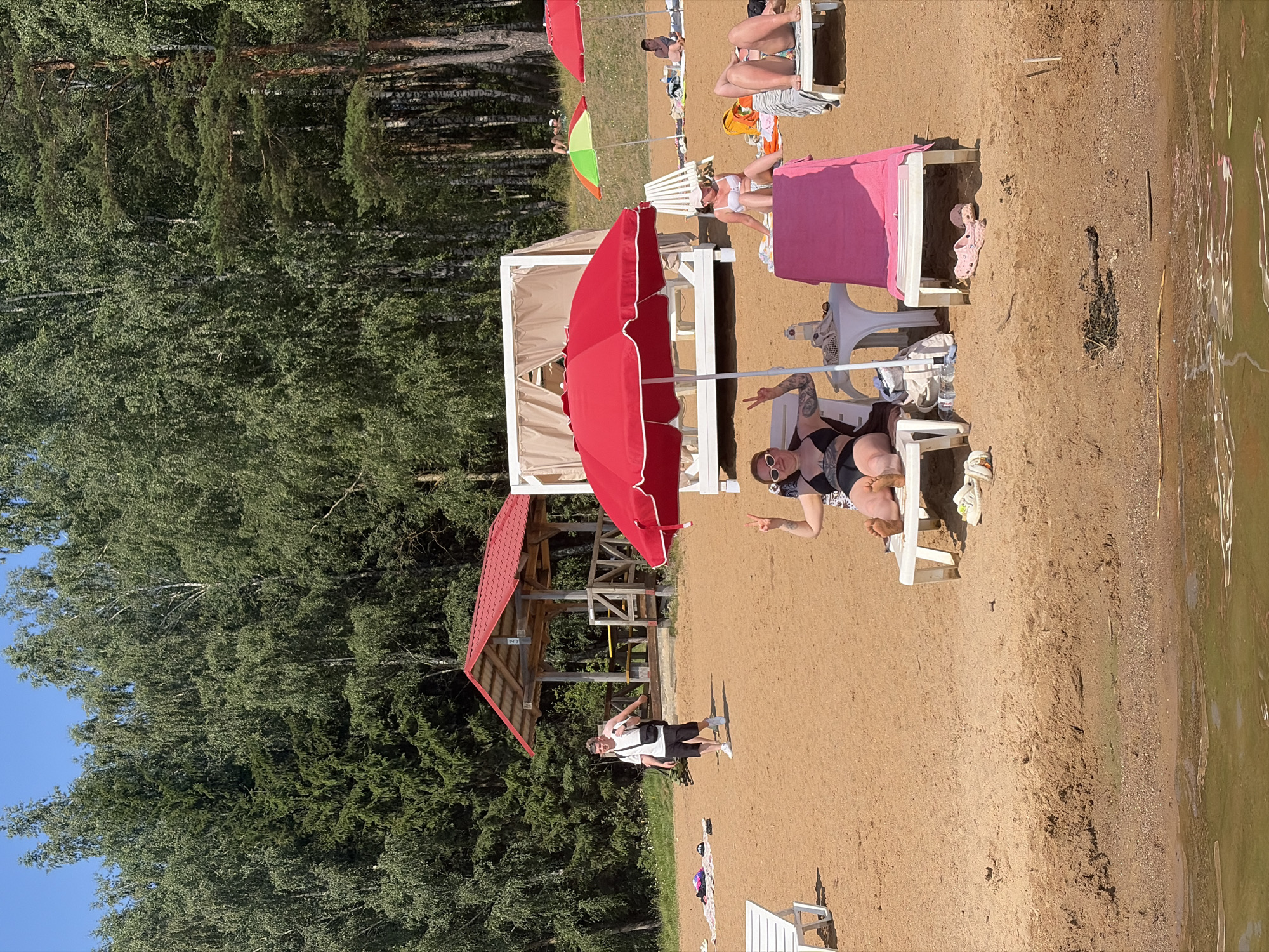 The image depicts a sunny day at a sandy beach, likely in Ozyaritska-Slabadski rural council, Belarus, given the location context. The scene is a summer leisure setting with several people relaxing on beach chairs. A prominent red umbrella shades a section of the sand, and a smaller, colorful umbrella is also visible. Two white canopies, possibly part of a beach club or relaxation area, are set up on the sand. In the background, a dense forest of green trees borders the beach, with a clear blue sky above.
Several individuals are captured in various states of relaxation. One person is lying on a beach chair in a black bikini, striking a peace sign with their fingers and wearing sunglasses. Another person in a white bikini is sitting on a beach chair, seemingly posing. Further back, other beachgoers can be seen lounging on chairs, some partially obscured by umbrellas or other beach furniture. A young man in a white t-shirt and black shorts is standing on the sand, looking towards the camera. The overall mood appears to be relaxed and enjoyable, consistent with a summer vacation or day at the beach. The lighting suggests it is midday, with bright sunlight casting shadows on the sand. The water of a lake or river is visible on the right side of the image, reflecting the sunlight and surrounding greenery. There is no visible text in the image.