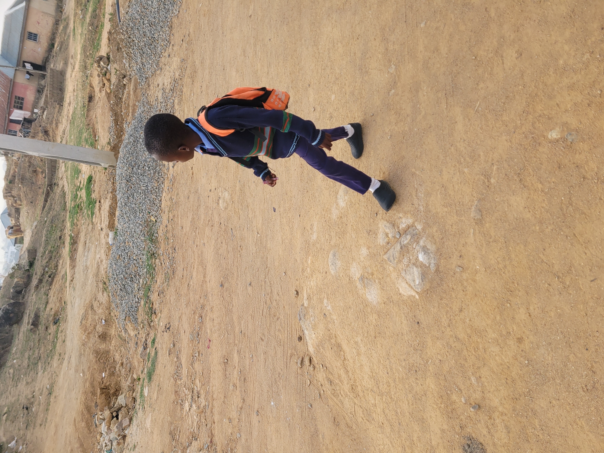 A young boy in a school uniform and orange backpack is running on a dirt path in Jos, Nigeria. The background includes a building, gravel, and vegetation.