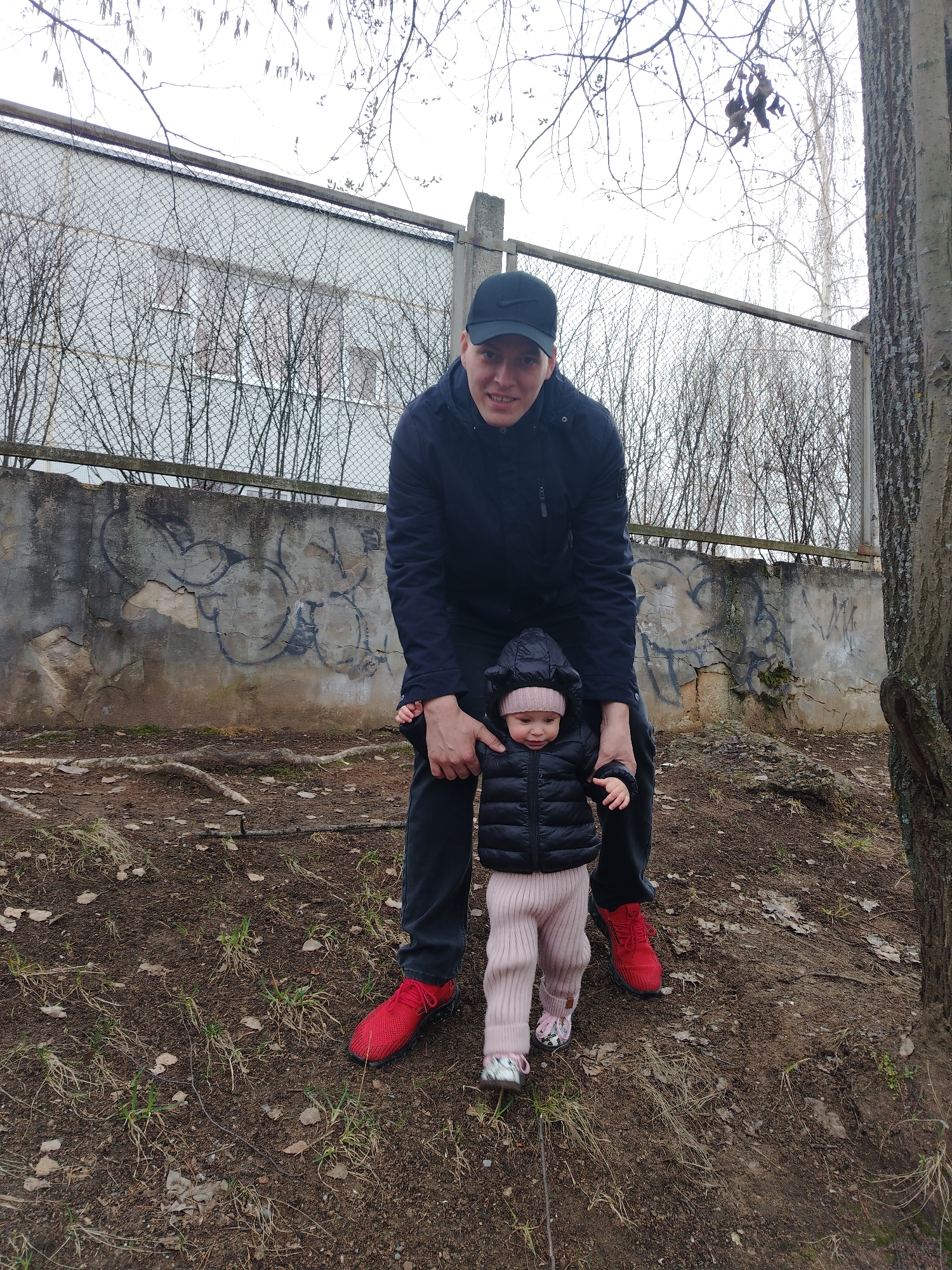 The image depicts an adult man and a young child outdoors, likely in Chaikovsky, Russia, on an overcast day. The man, wearing a black baseball cap with a white logo (resembling a Nike swoosh), a black puffer jacket, dark trousers, and bright red athletic shoes, is smiling towards the camera. He is holding the hands of a small child, supporting them as they stand or walk.
The child is dressed in a black puffer jacket, a pink knitted hat with small ear-like details, light pink ribbed pants, and patterned shoes. The child is looking down and to the side, appearing focused.
They are positioned on natural ground covered with dirt, sparse dry grass, and exposed tree roots. In the background, a weathered concrete wall, adorned with unreadable black graffiti, is topped by a chain-link fence. Behind the fence, bare tree branches and a light-colored building with windows are visible. A large tree trunk is prominent on the right side of the frame. The bare trees and the subjects' warm clothing suggest a cool season, such as late autumn or early spring. The scene conveys a tender interaction as the adult assists the child.