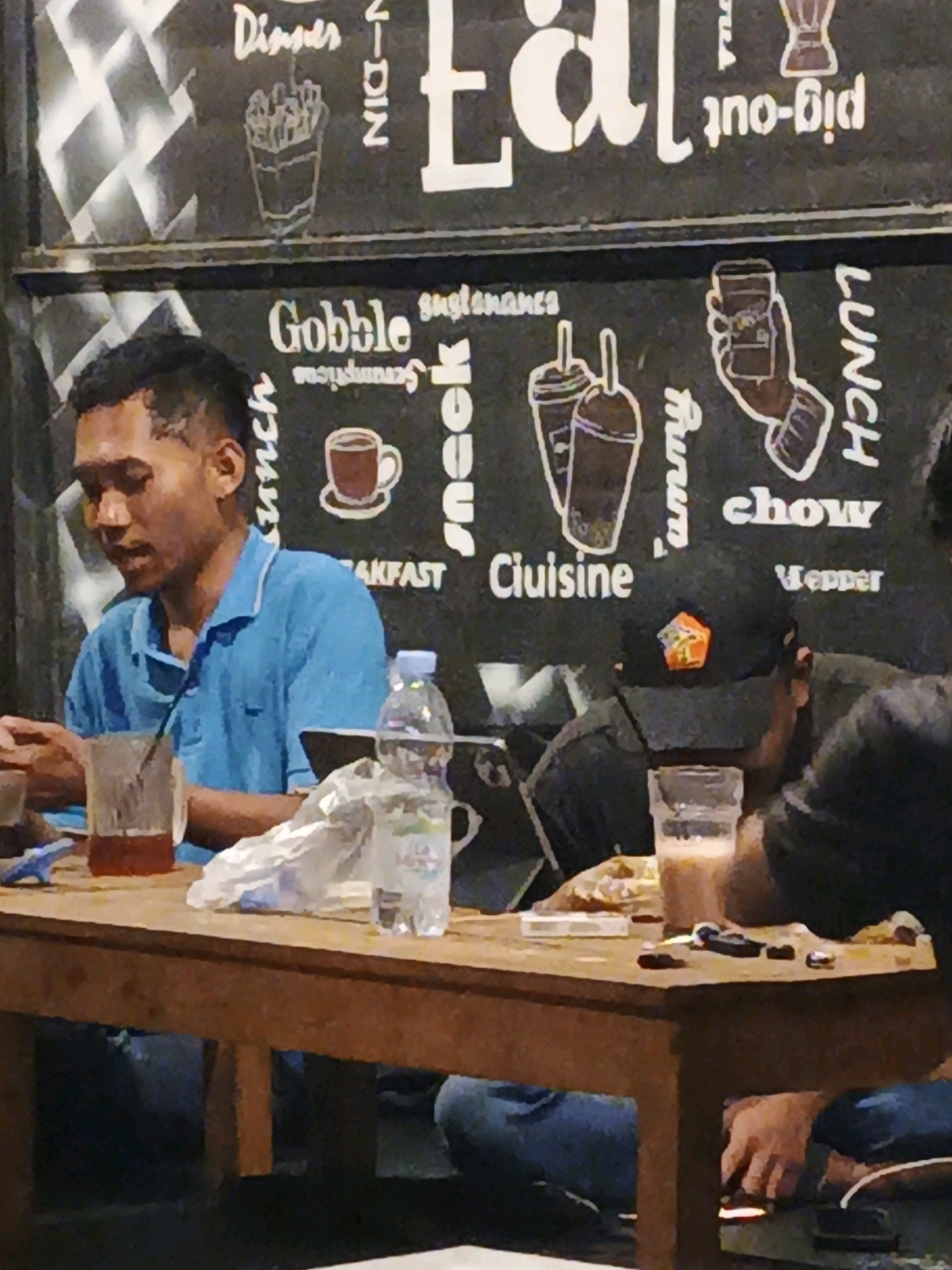 In Ketegan, Indonesia, a group of individuals are gathered around a wooden table, engaged in a meal. A man in a blue polo shirt is seated on the left, looking to his left while holding a glass of amber liquid with a straw. On the table in front of him, a crumpled plastic bag and a water bottle are visible. To his right, another person wearing a black baseball cap with an orange emblem is seated, leaning forward and holding a glass of a light brown beverage. The background features a black wall with white text and illustrations, including stylized depictions of food and drinks, and words like "Dinner," "Eat," "Gobble," "Snack," "Lunch," and "Cuisine." The lighting in the scene is dim, creating a relaxed ambiance.