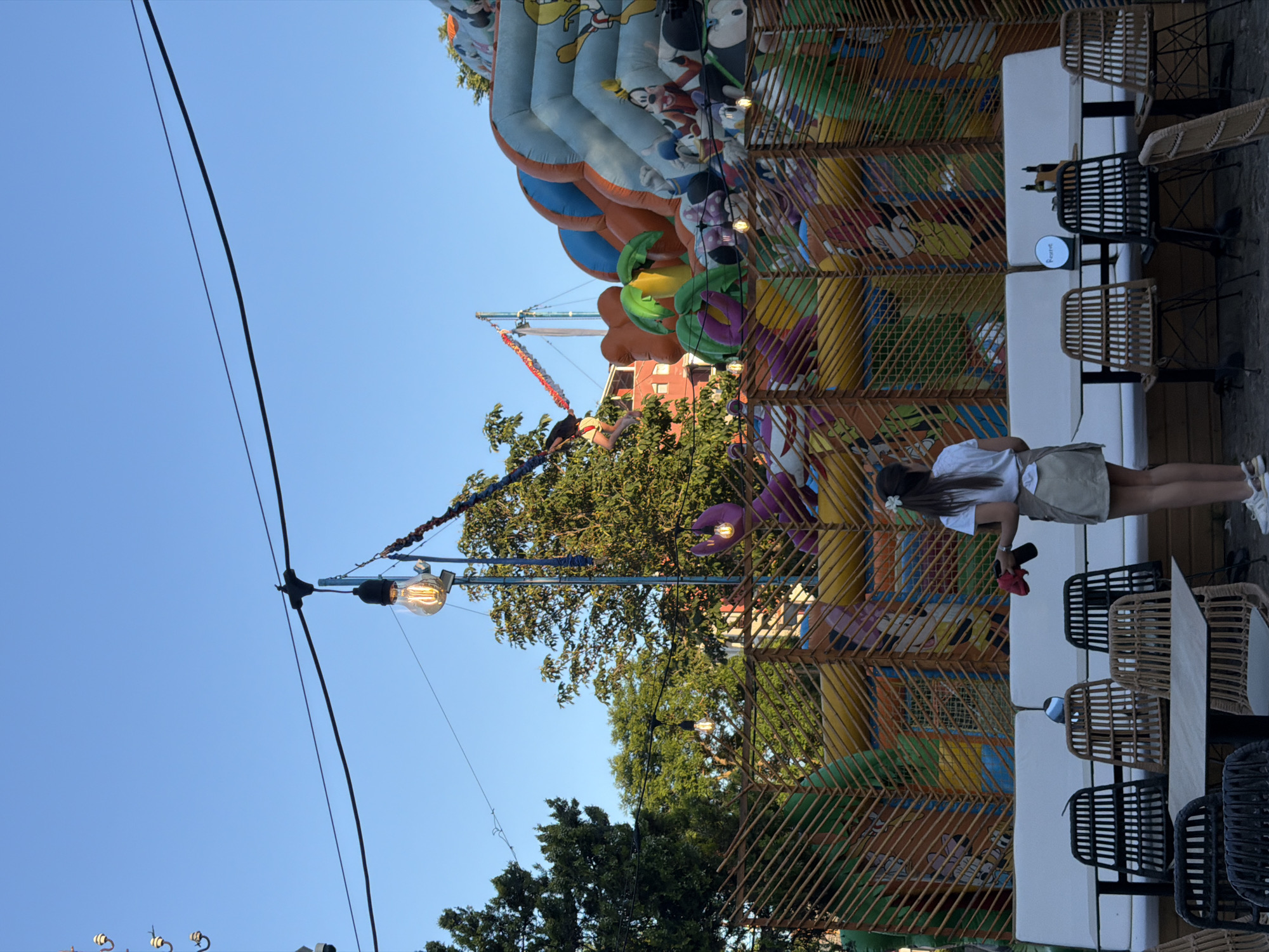 In the city of Sozopol, Bulgaria, a woman stands facing away from the camera, holding a red cloth in her right hand. She is wearing a white t-shirt and a beige skirt. The woman is positioned in front of a white bench and several chairs surrounding tables. Behind the bench, a large inflatable structure, decorated with cartoon characters like Mickey Mouse and Donald Duck, is visible. The inflatable structure appears to be a bouncy castle or slide, with various colorful elements and climbing sections. To the left of the inflatable, a young person is suspended in the air, attached to a bungee cord. The background shows trees and a clear blue sky. Overhead, string lights with exposed bulbs are strung across the scene, suggesting an outdoor entertainment or dining area. The overall atmosphere appears to be that of a recreational park or amusement area.
