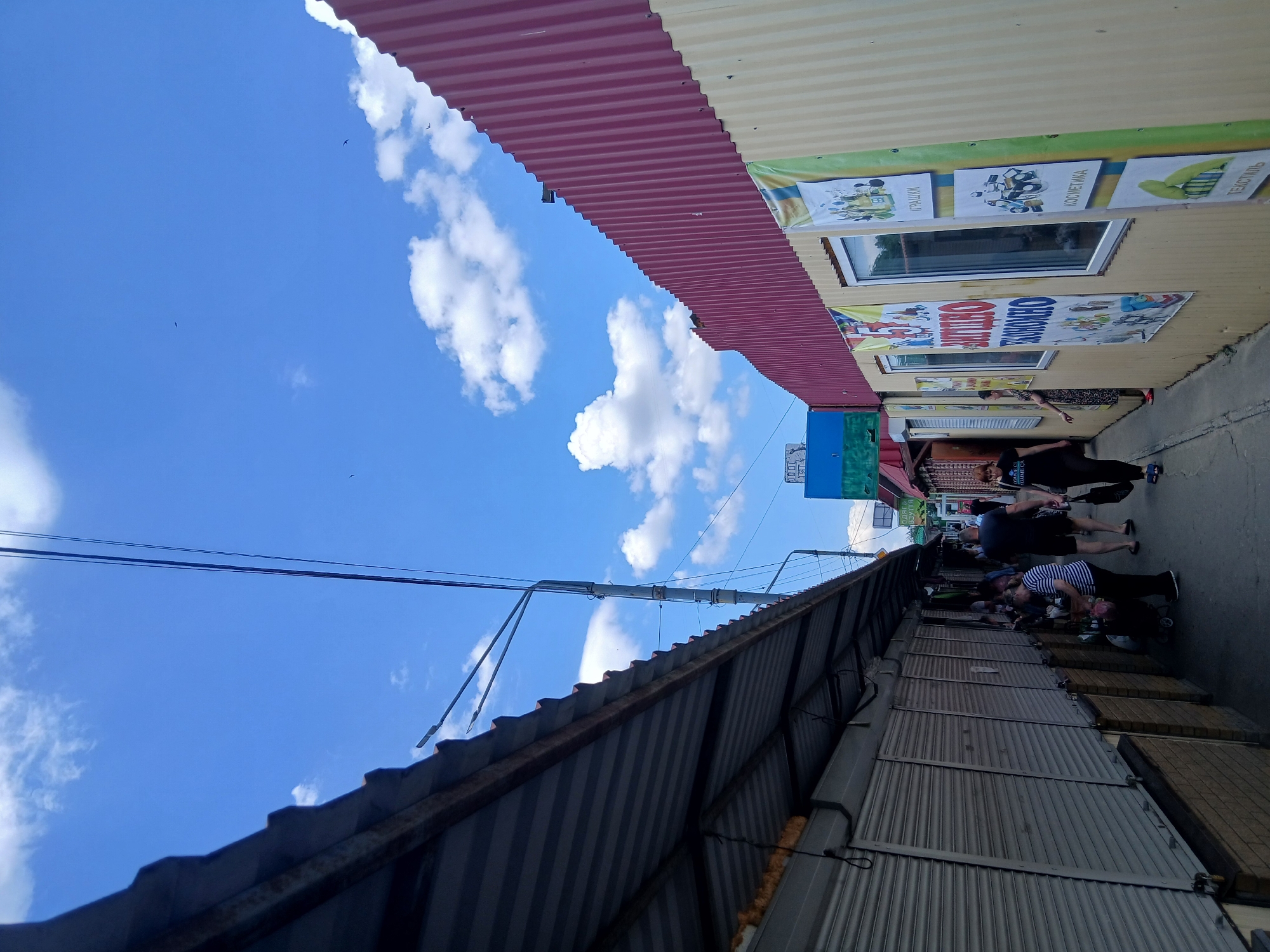 In Dnipro, Ukraine, a narrow market alleyway is depicted under a bright blue sky with scattered white clouds. On the left, a row of closed market stalls with corrugated metal shutters lines the path. A corrugated metal roof extends over these stalls. On the right, a building with a red corrugated metal roof has a yellow facade with posters. One poster is large and red with Cyrillic text, and other posters depict various items like toys, cosmetics, and textiles. Several people are walking in the alley. A man and a woman are walking away from the camera, with the woman in a black t-shirt and dark pants, and the man in a dark t-shirt and shorts. Another woman in a striped shirt is seen with a shopping cart. The ground is paved. The scene appears to be a typical outdoor market environment.