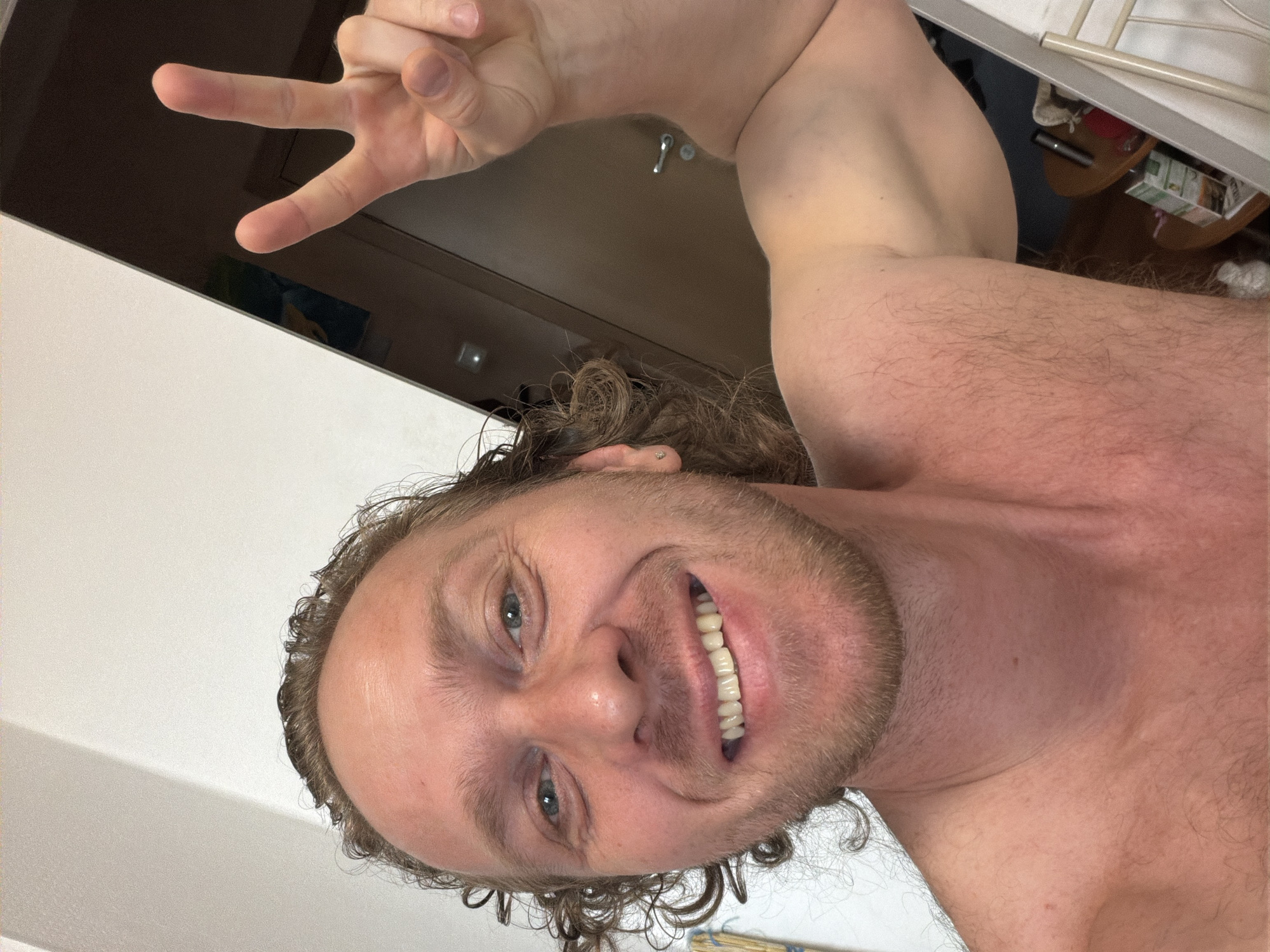 A selfie of a man with fair skin, light brown hair with some curls, and a beard, smiling with his mouth open, showing his teeth. He is making a peace sign with his left hand. The background appears to be a simple interior with a door. The location cannot be determined from the image.
