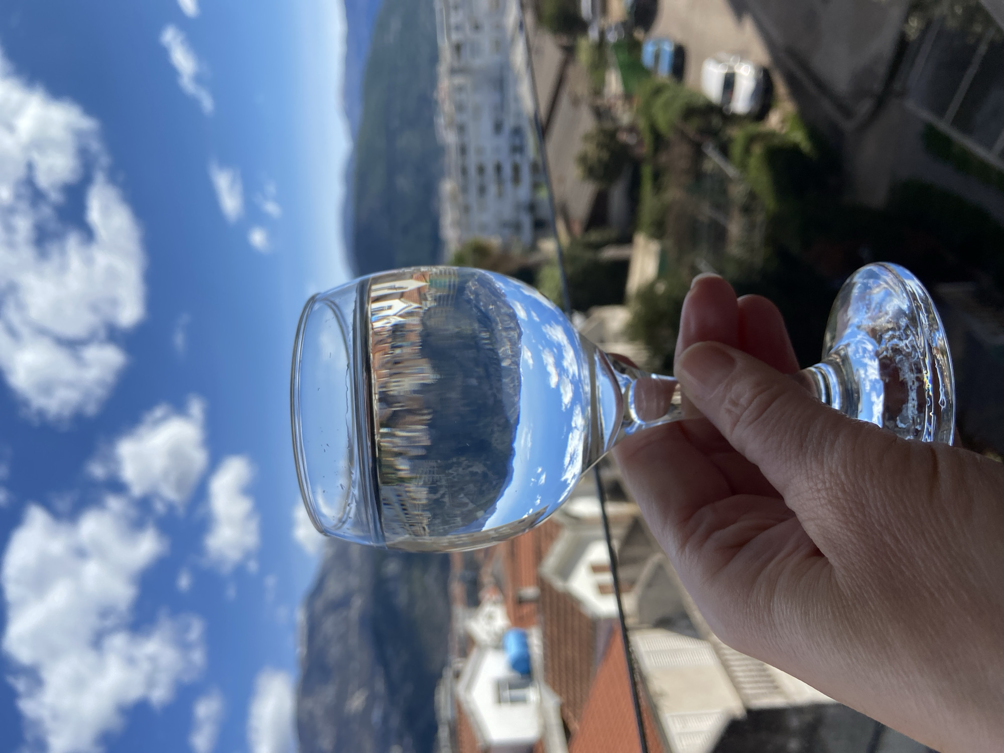 A hand holds a glass of clear liquid, reflecting the surrounding landscape upside down. In the reflection, one can discern mountains, buildings, and a blue sky with white clouds. The background, outside of the glass, shows the city of Budva, Montenegro. Visible are rooftops of houses, more buildings, and lush greenery. The sky is visible above the glass, mirroring the scene inside.