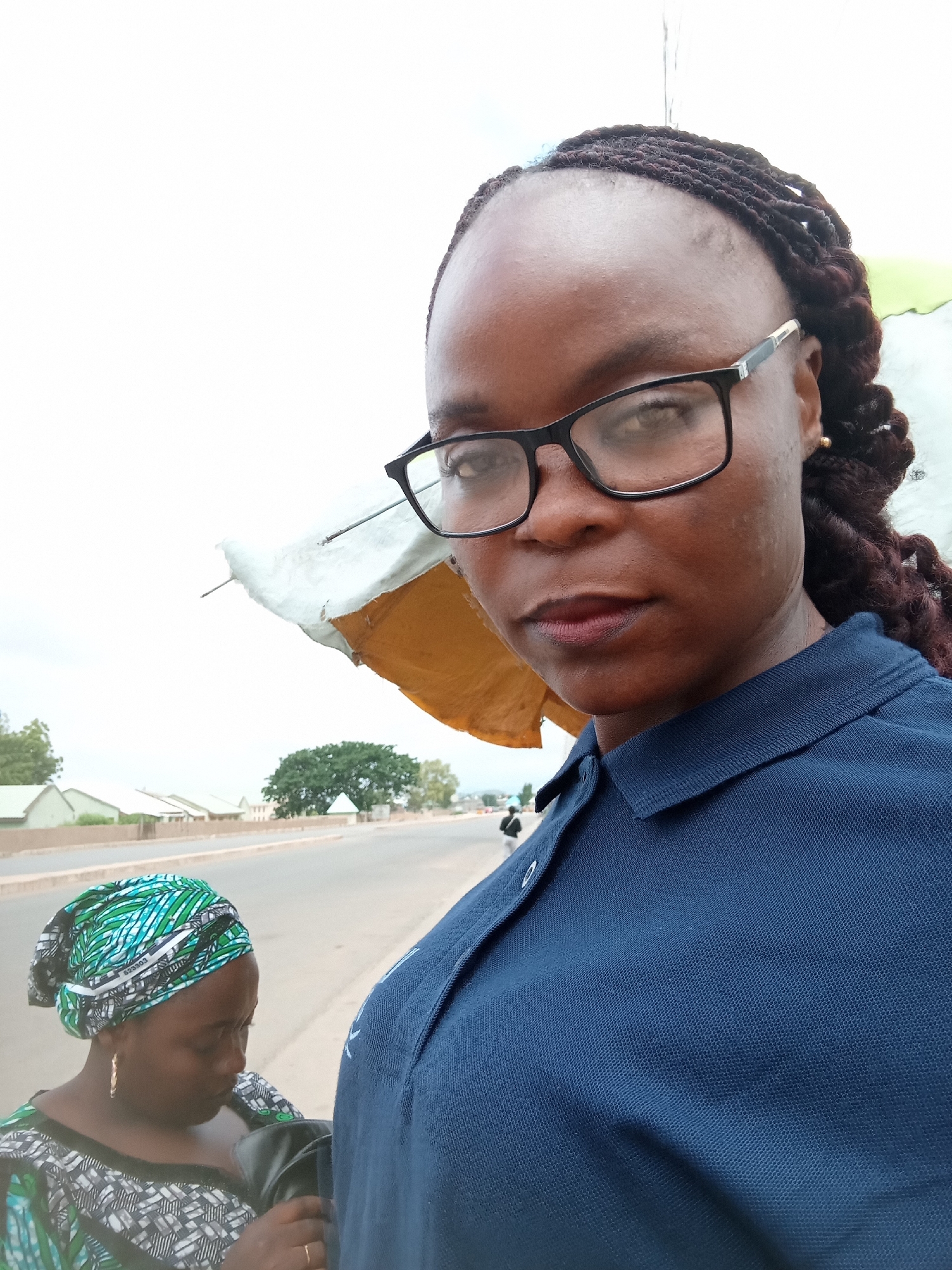 In an outdoor, urban setting, two women are visible. The primary subject, a woman wearing black-rimmed glasses and a dark blue polo shirt, is positioned in the foreground, facing the camera. Her hair is braided and pulled back. To her left and slightly behind her, another woman is visible, wearing a patterned headscarf in green and white, and a black and white patterned garment. She appears to be looking down. In the background, a road runs horizontally, with buildings and trees lining its sides. A person is walking on the road in the distance. A portion of a white and yellow umbrella structure is visible near the primary subject, obscuring part of the background. The sky is overcast.
