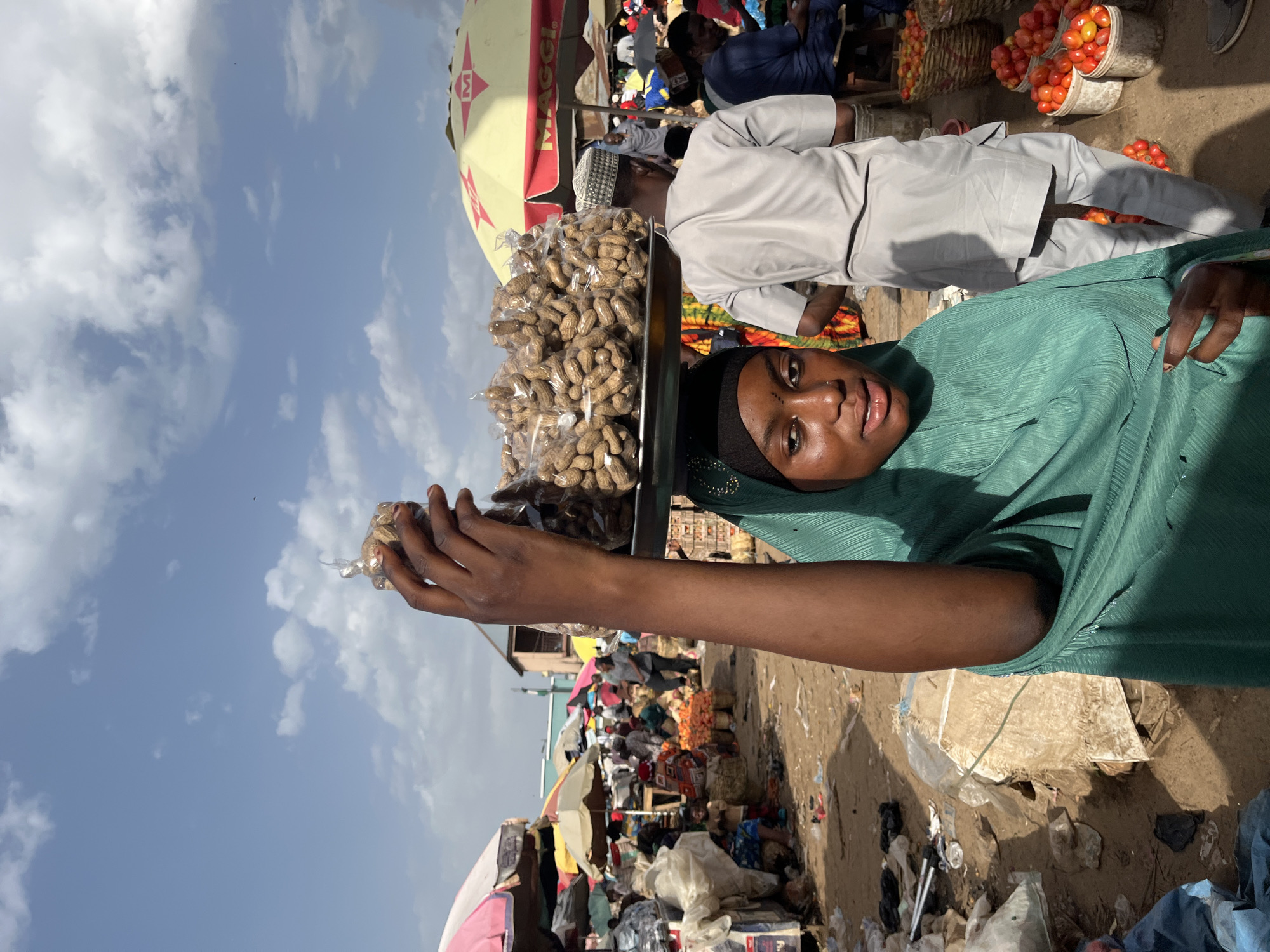 A young Black woman wearing a teal hijab stands in a bustling outdoor market, balancing a large tray of peanuts in plastic bags on her head. Her face is turned towards the camera, and she has a slight smile. Behind her, a man in a light gray traditional tunic and cap walks away from the camera, and further in the background, various market stalls with umbrellas and baskets of produce are visible. The ground is dirt and littered with some trash, suggesting a typical street market environment. The sky above is blue with scattered white clouds. The location is the city of Rafin Par, Nigeria.