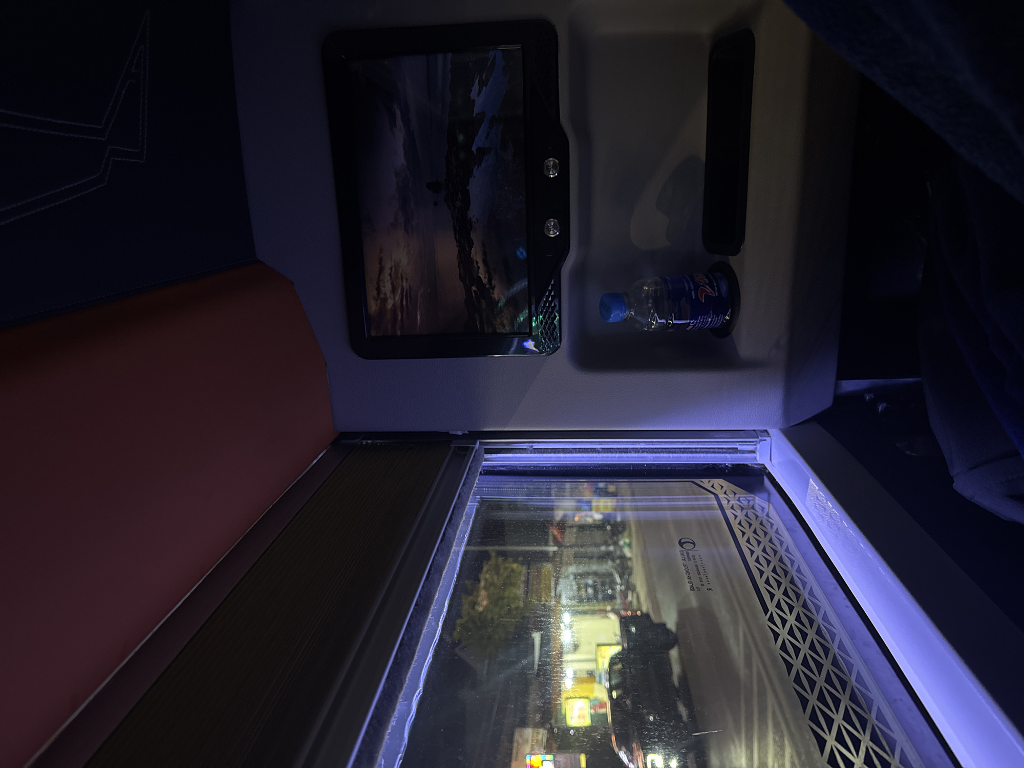 The image shows an interior view from a reclining seat, likely inside a bus or train, during an evening or nighttime setting in Phi Nôm, Vietnam. The window reflects the illuminated street scene outside. A personal entertainment screen is mounted on the wall in front of the seat, displaying an image. A bottle of water is placed in a cup holder nearby. A partially drawn window shade is present. There are no people visible, only an unseen passenger's blanket.