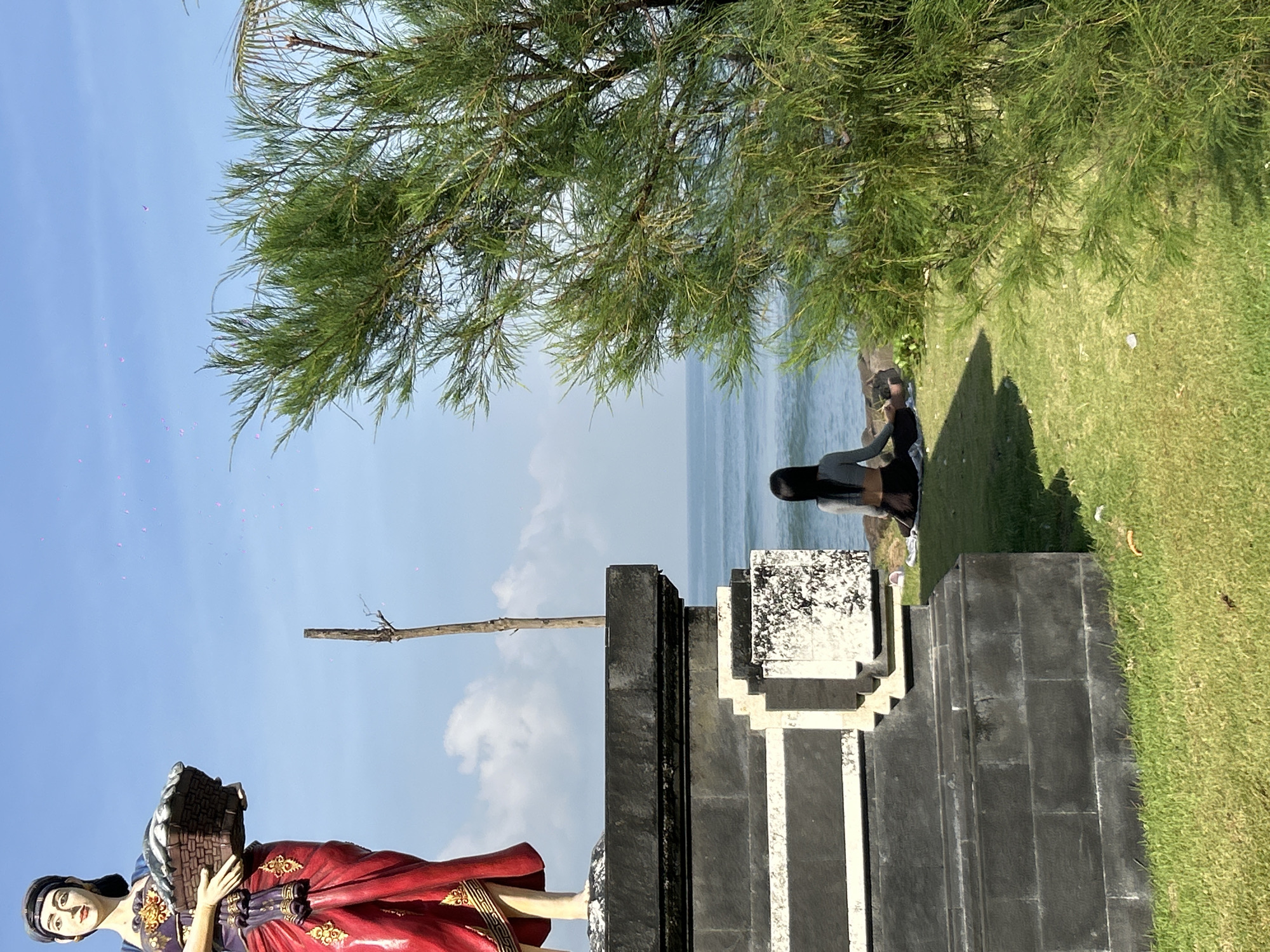 Captured at an extreme angle, the scene presents a confluence of elements from Mengwi, Indonesia. A statue of a figure holding a basket stands prominently, juxtaposed against a structure that appears to be a stone building. A woman is sitting near the structure, gazing out toward the water. Lush green foliage, including what seems to be a tree, occupies the top portion of the frame. The sky is clear blue, with a few light clouds.