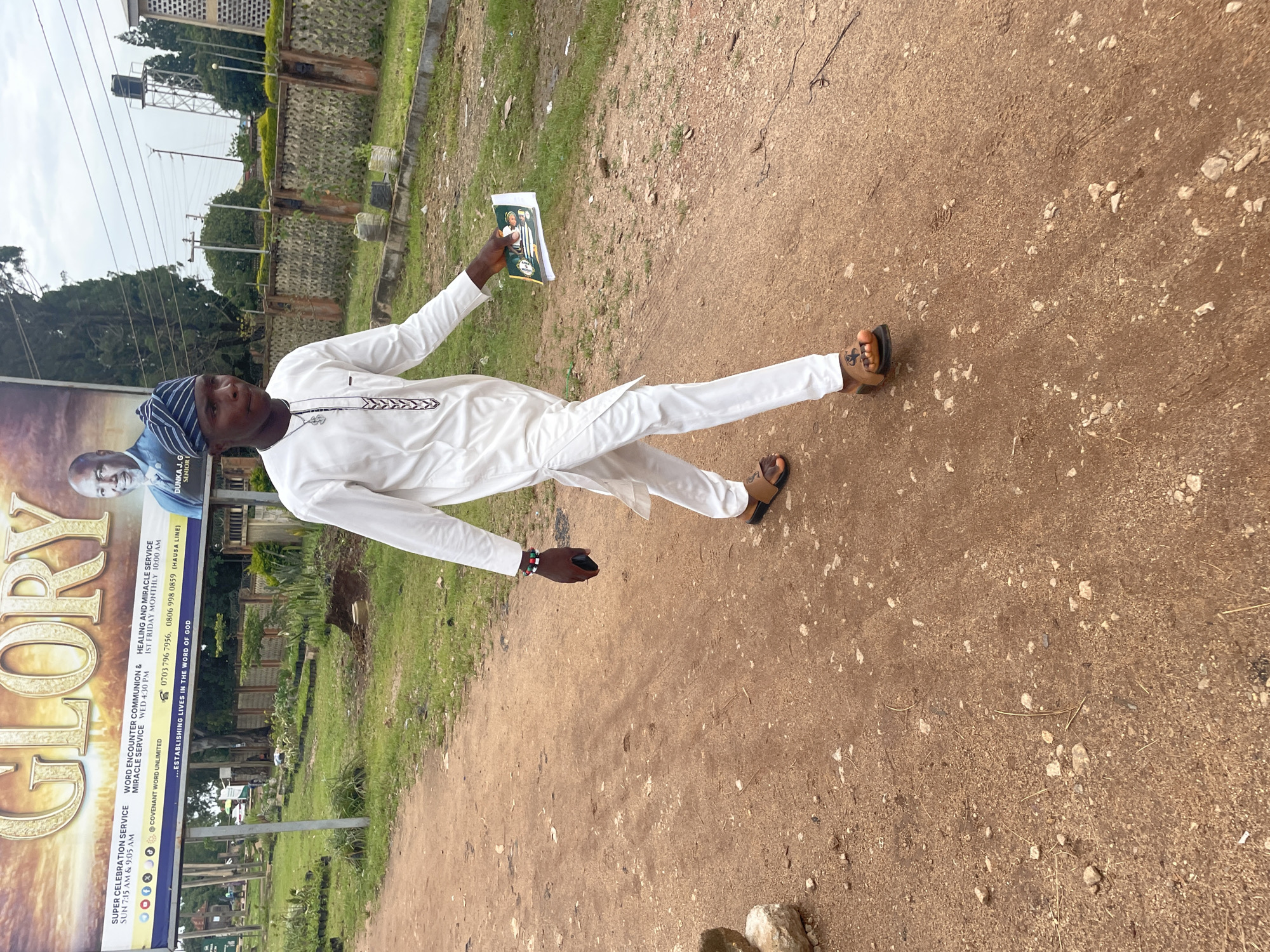 A young man walks on a dirt path in front of a billboard in Jos, Nigeria. He is wearing a white traditional outfit and a striped head covering, and he is holding a pamphlet. The billboard advertises a church service and features a picture of a pastor. The path is lined with green grass and small trees. In the background, there is a building with a water tower and a fence. The sky is overcast.
