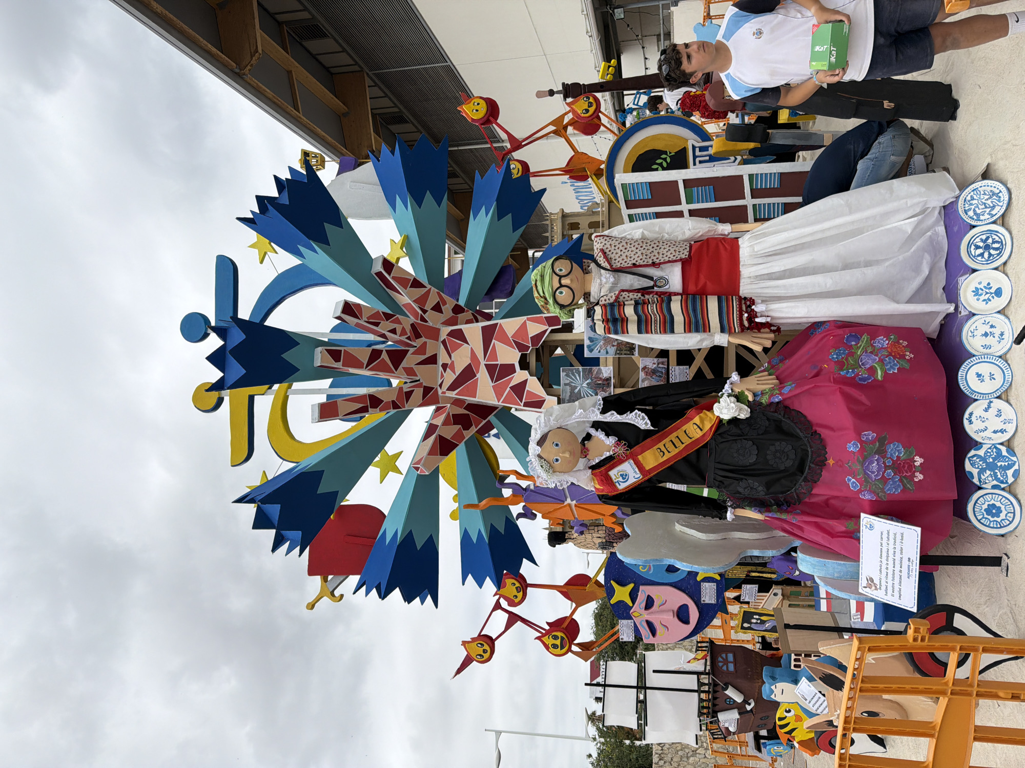 The image shows a street scene in an outdoor environment. In the foreground, there are two large, standing figures resembling mannequins, dressed in traditional attire. The figure on the left wears a black dress with a floral pattern on the skirt and a red sash that reads "BELLEA". The figure on the right wears a white dress, a red sash, a patterned vest, a striped scarf, and glasses. Behind them is a large, colorful structure that appears to be a parade float or monument, featuring geometric shapes, stylized figures, and a Ferris wheel-like element. The background includes buildings, a cloudy sky, and a young man in a t-shirt and shorts standing on the right side of the frame, holding a green box. Several decorative plates are arranged in front of the figures on the ground. The scene appears to be set up for a festival or public event.