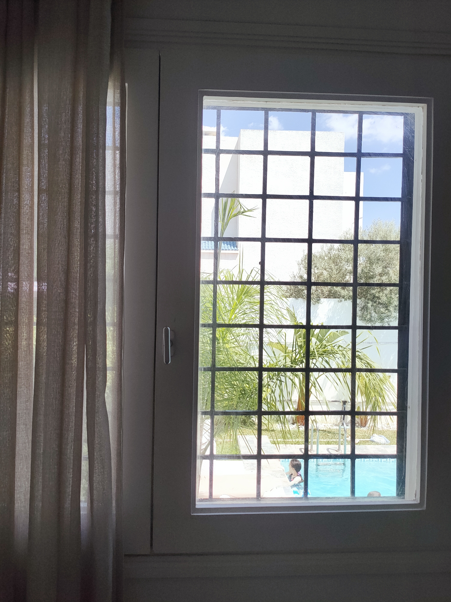The image captures a view through a window with a black metal grate, looking out onto a courtyard with a swimming pool. To the left, a beige curtain is drawn partially, revealing a second window behind it. The main view through the grates shows a bright blue swimming pool with a ladder. Two children are partially visible in the pool; one child is near the ladder, and another, further to the right, is only their head visible. In the background, there is a white building and green palm trees, indicative of a sunny, possibly Mediterranean climate. The scene appears to be a residential or resort property. Based on the architectural style and vegetation, the location is likely Mrezga, Tunisia, known for its coastal resorts and white-washed buildings.