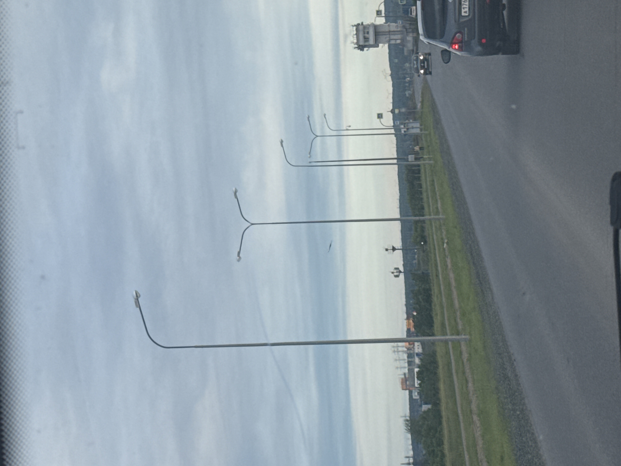 The image shows a view from inside a car traveling on a highway. Several streetlights line the road with cloudy skies above. To the right, a grassy median divides the highway. Cars can be seen ahead, including a dark-colored vehicle with the license plate "A176". In the distance, buildings can be seen. Based on the surroundings, the location is identified as Chaikovsky, Russia.