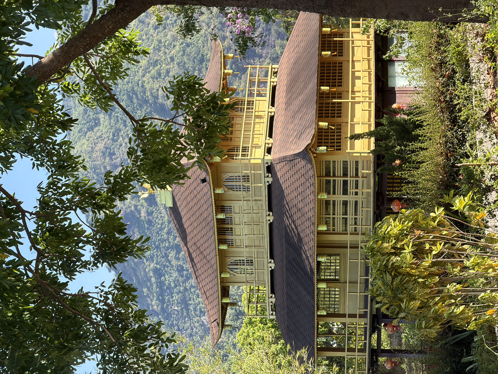 A tall, yellow, pagoda-style building with a dark brown roof stands against a backdrop of lush, green mountains and a clear, blue sky. The building has multiple levels with intricate window patterns and decorative elements hanging from the eaves. Part of the building is obscured by dense foliage, including trees with bright green leaves in the foreground and a mix of shrubbery with red lanterns near the base. The scene is likely located in or near a mountainous area, possibly in Nam Nha Trang Ward, Vietnam, given the presence of tropical vegetation.