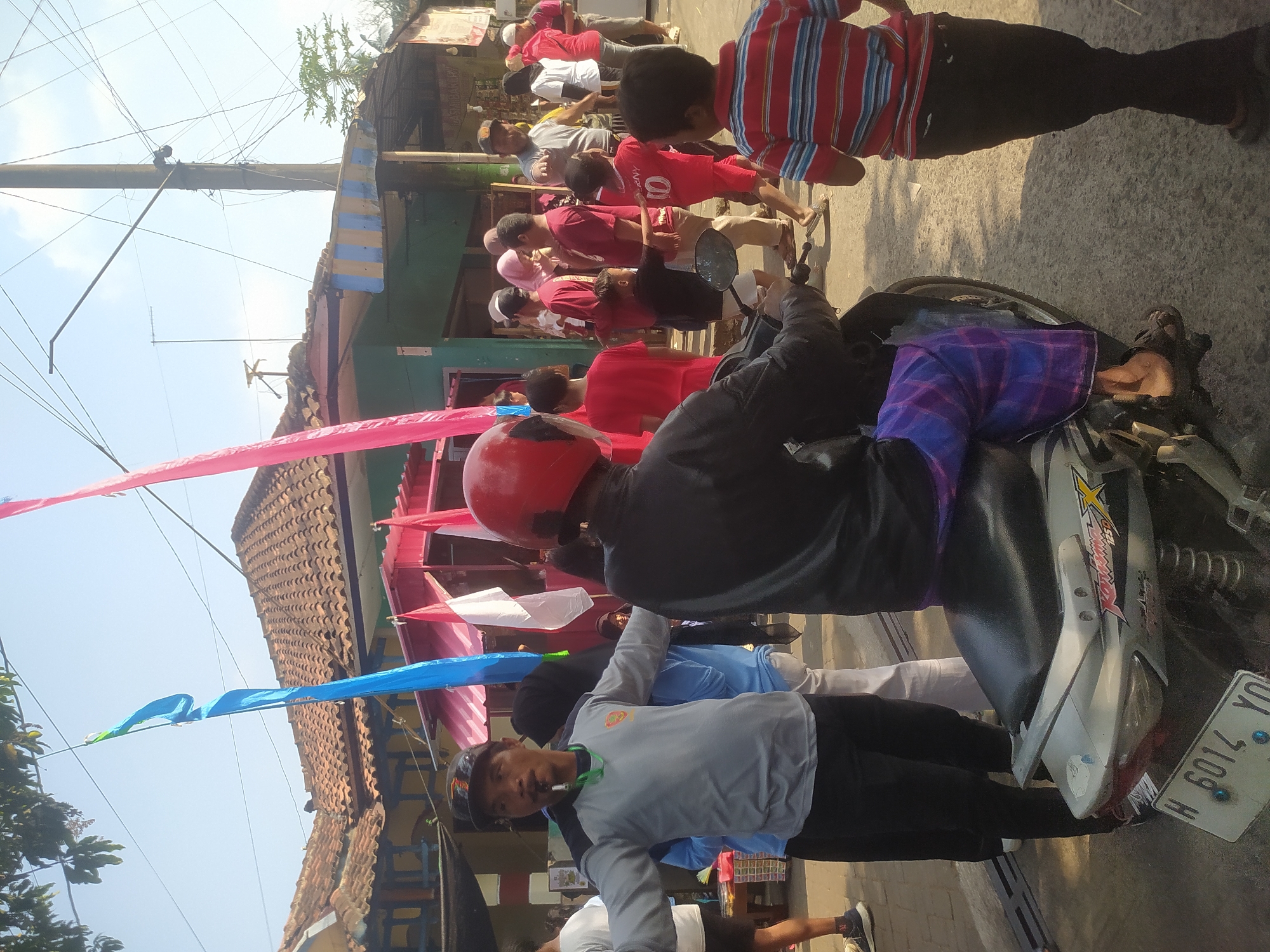 In Lopait, Indonesia, a man in a gray shirt and black pants, wearing a red helmet and a blue and purple checkered sarong, sits on a silver motorcycle. He is holding a whistle in his mouth and has his arms outstretched. A group of people, some wearing red shirts, are gathered behind him. Two flags, one blue and one pink, are flying in the background. The scene appears to be a street or an outdoor gathering area, with buildings and power lines visible.