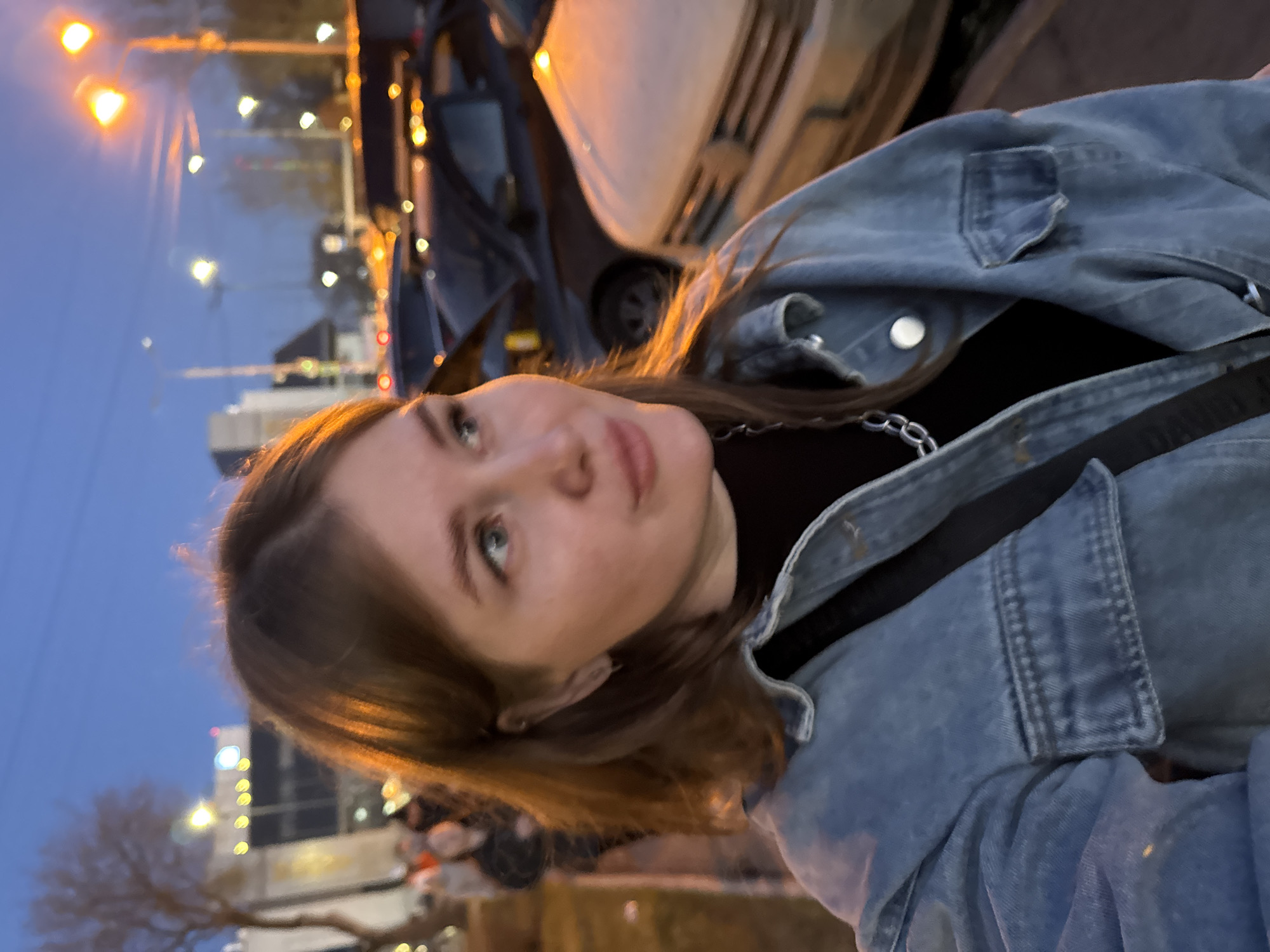 The image depicts a young woman in a denim jacket, standing outdoors at dusk in what appears to be an urban setting.
The woman has fair skin, brown hair, and light-colored eyes. She is wearing a black top underneath the jacket, with a chain visible around her neck. A strap is visible across her chest.
The background shows city lights and buildings. A car is parked nearby, partially obscuring the view. The lighting suggests it is early evening, with streetlights illuminating the scene. Bare branches of trees are visible.
The location is identified as Perm, Russia, based on available information.