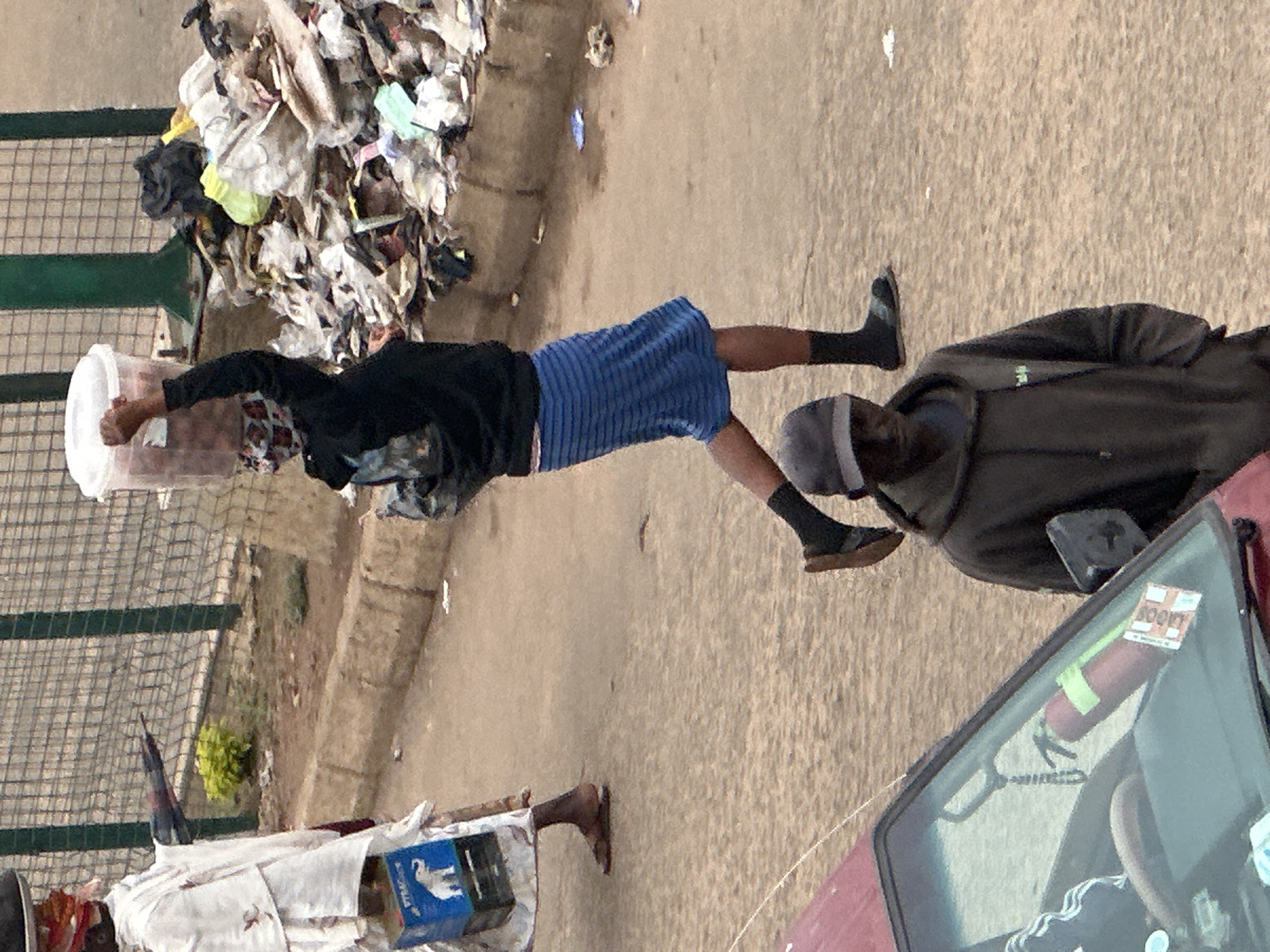 The image depicts an outdoor scene in Bukuru, Nigeria, characterized by a street, a roadside with a fence and debris, and several individuals. In the foreground, a man wearing a dark gray hooded sweatshirt and a baseball cap is standing, facing towards the camera. To the left of the frame, a portion of a red car's windshield is visible, with a fire extinguisher mounted on it and a sticker attached to the windshield.
Further back, a woman is walking, carrying a large clear plastic container filled with what appears to be food items on her head. She is wearing a black hooded sweatshirt and a blue and white striped skirt, along with dark socks and sandals. To her left, another person in white clothing is partially visible, carrying a blue box with an elephant logo.
The background features a chain-link fence bordering a raised area with some greenery and a large pile of garbage, primarily composed of plastic bags and assorted waste. The ground surface is a dusty, sandy road. The lighting suggests an overcast day.