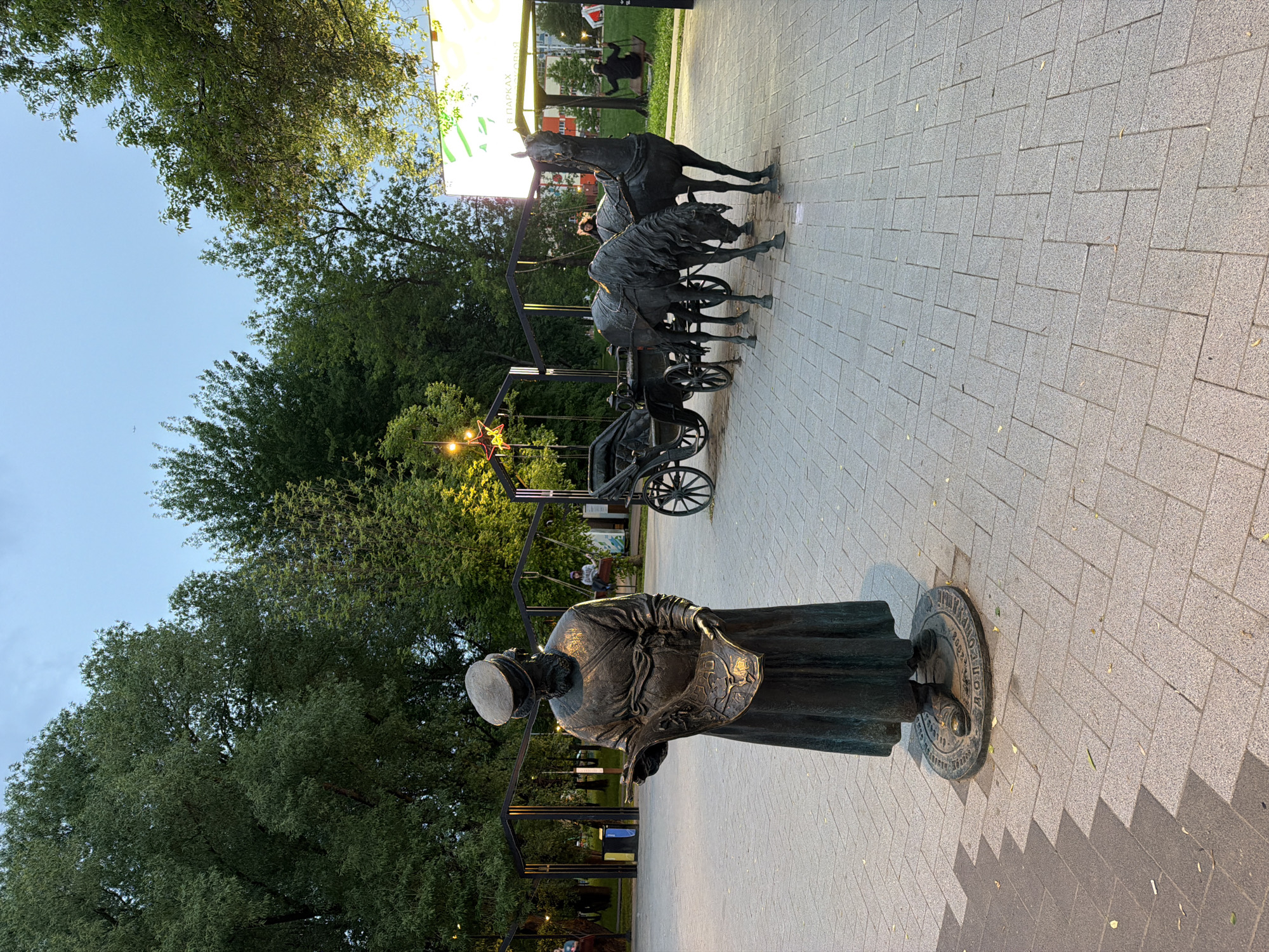 The image shows a bronze statue of a woman in a long dress and head covering, standing on a circular base. She is holding a bag. Behind her, there are bronze statues of horses and carriages. These statues are located on a paved area with trees and streetlights in the background. The location is Dolgoprudny, Russia.
