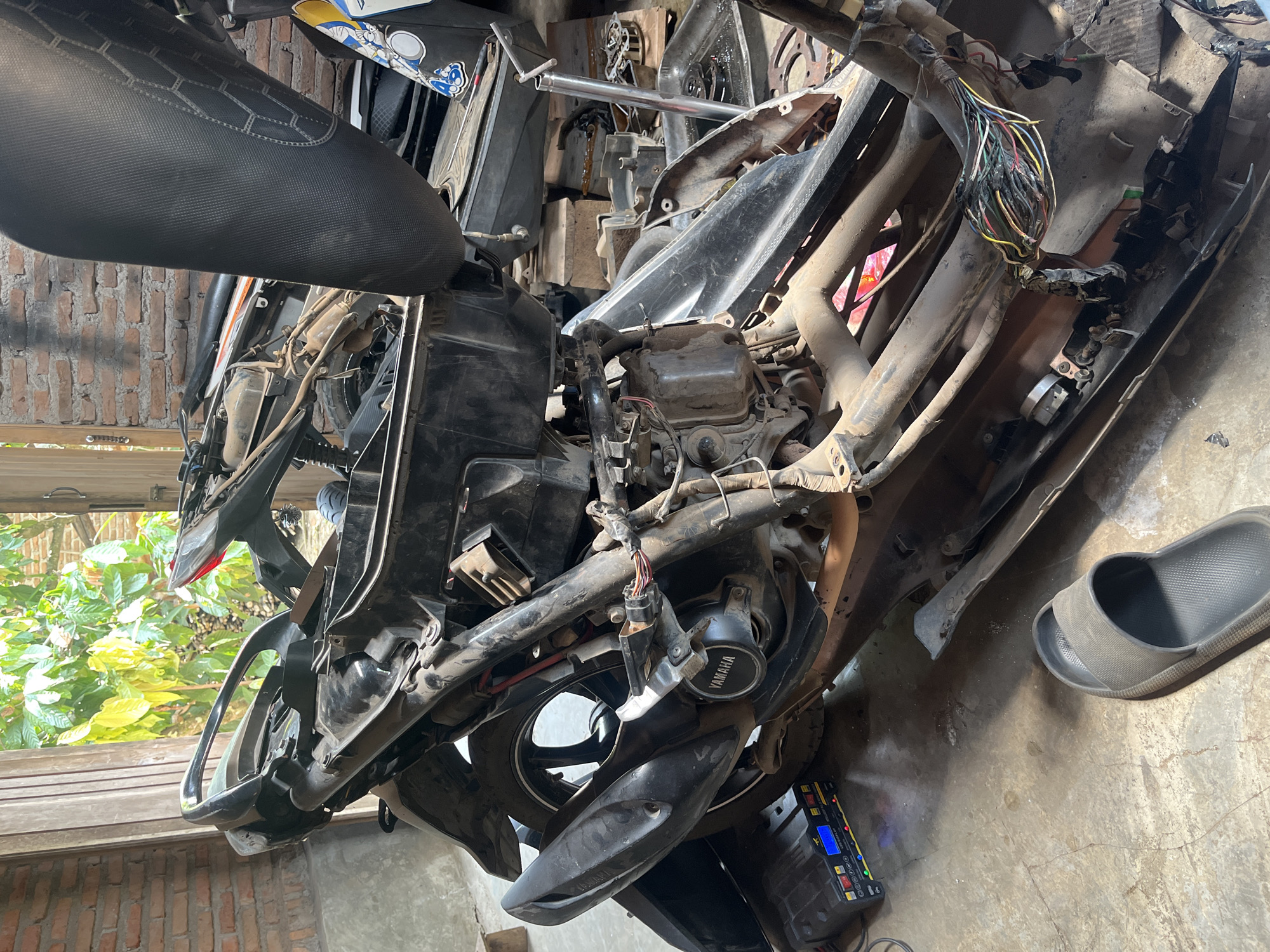 The image shows a partially disassembled black motorcycle in what appears to be a garage or workshop in Kebumen, Indonesia. The motorcycle is in a state of repair or maintenance, with various parts removed and wires exposed. A black flip-flop sandal is visible on the concrete floor. In the background, a brick wall and an open doorway revealing greenery suggest an outdoor area. A battery charger is positioned near the motorcycle. The overall scene depicts a utilitarian environment focused on vehicle repair.