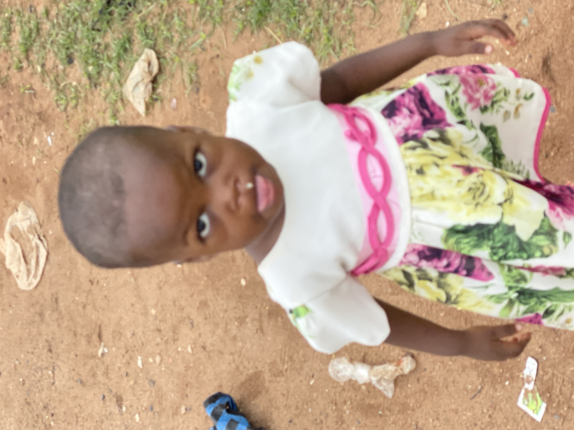 A young child stands on a dirt surface in Nkpor, Nigeria. The child wears a light-colored dress with pink and floral accents. The ground is littered with small debris and trash. A blue and black object, possibly a shoe, is at the bottom left of the frame. The child is looking up and towards the camera with its tongue slightly protruding.