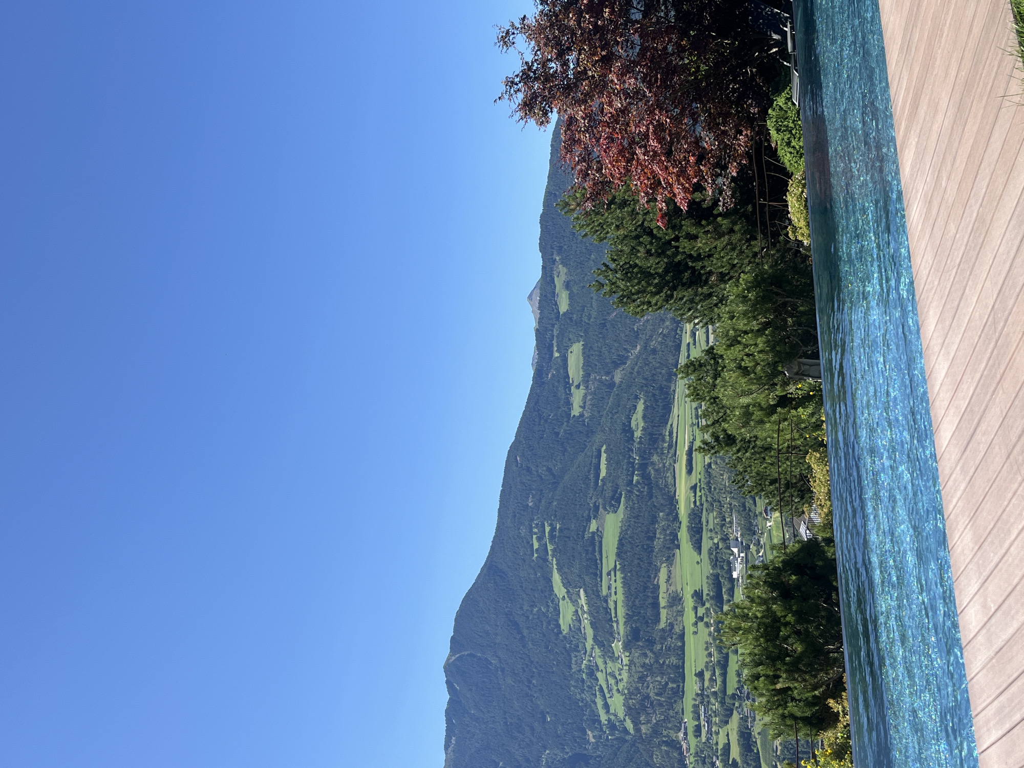 The image depicts an infinity pool in the foreground with a wooden deck. Beyond the pool, a lush green landscape with a village nestled in the valley unfolds, leading to a mountain range covered in trees. The sky is a clear, vibrant blue. This scene is located in the vicinity of Brixen - Bressanone, Italy, a city known for its alpine setting.
