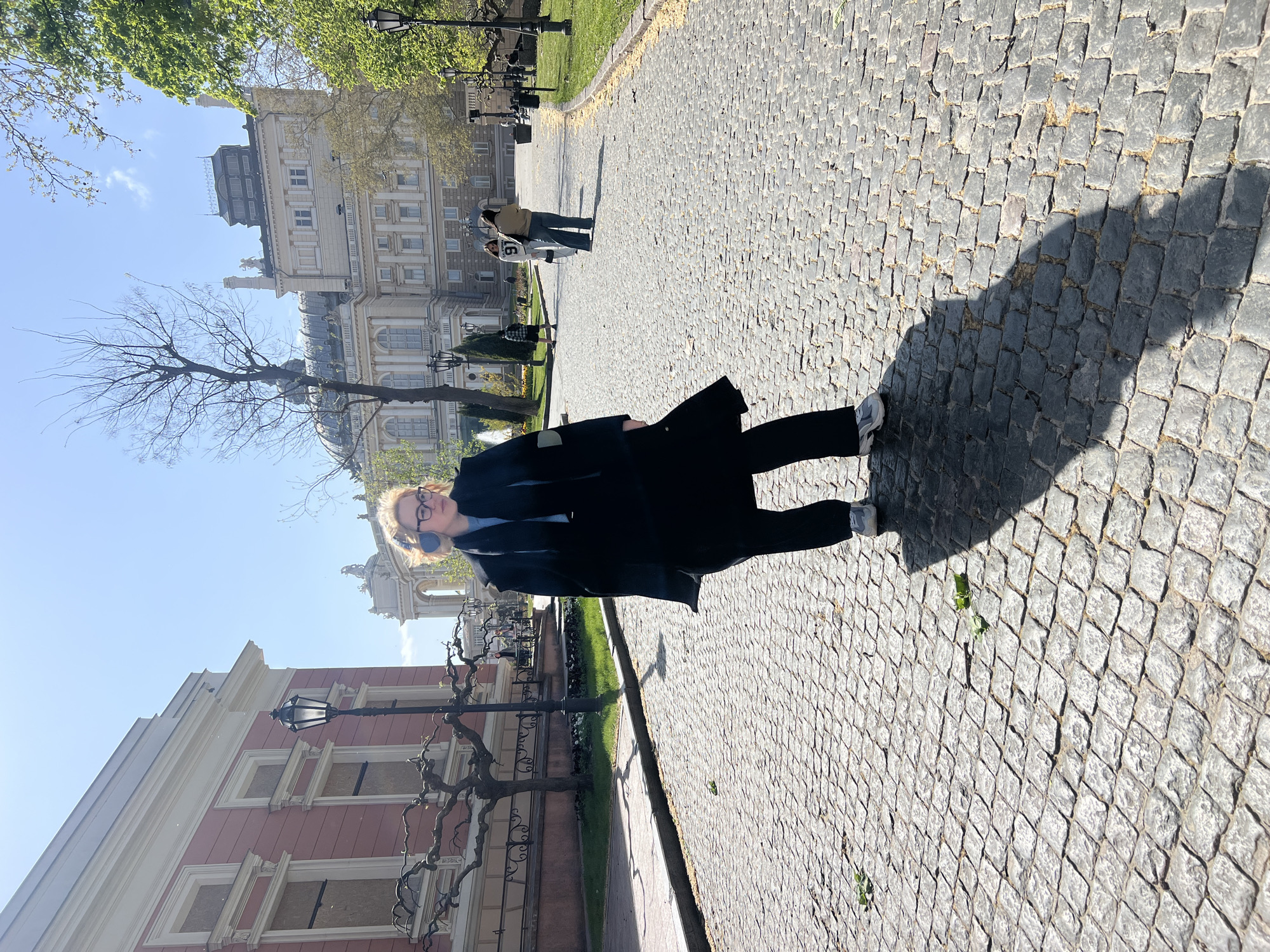 A full-body shot depicts a woman wearing glasses, headphones, a black coat, dark pants, and white shoes. She stands on a stone-paved walkway. Her shadow stretches to the right, indicating a strong light source to the left. In the background, buildings can be seen. The image appears to have been taken in Odesa, Ukraine, as evidenced by the architecture and the paving stones. Specifically, the architecture suggests the photo was taken in a central, historic part of the city. Other individuals are visible farther down the path. The environment suggests a park or square in the city. The trees are bare except for the trees at the top left of the image. A red building with windows is visible on the left side of the image.