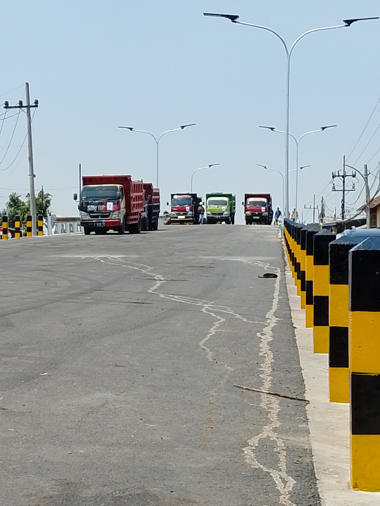 In Pasuruan, Indonesia, on a bright, clear day, several trucks are seen on a road, possibly a bridge. In the foreground, a gray asphalt road curves to the right, bordered by a white line and a series of black and yellow striped bollards. In the middle ground, a convoy of trucks moves away from the camera. The lead truck is red and white, with "Sempurna" written on its side. Behind it, a green truck and then another red truck follow. In the background, more trucks are visible, along with streetlights and utility poles. A few people are seen walking near the trucks. The environment appears to be an open area with a clear sky.