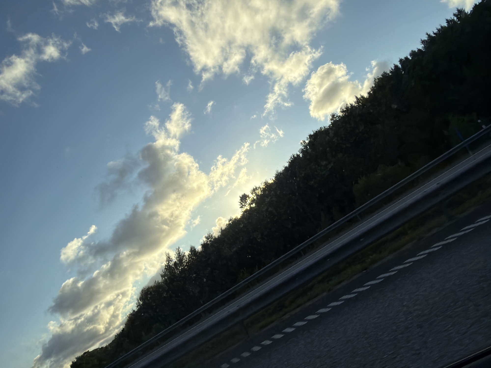 The image shows a tree-lined highway in Ödsmål, Sweden, under a bright blue sky with scattered white clouds. The sun is visible through a break in the clouds, casting a warm glow on the scene. The highway appears to be a multi-lane road with a guardrail separating the lanes. The trees are dense and dark, forming a thick forest along the side of the road. The ground is covered with asphalt, and there are white dashed lines marking the lanes. The image is taken from a slight angle, giving a sense of motion.