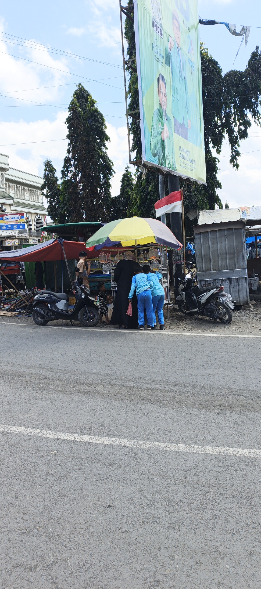This media file depicts a street scene in Puger, Indonesia, under a bright, partly cloudy sky, suggesting it's daytime. In the foreground, a grey asphalt road with a white line runs across the image. Behind the road, a bustling market stall is set up. Several people are present, including two individuals in bright blue outfits, one of whom is holding a red plastic bag, and another person in a brown outfit. A child in a patterned outfit is also visible.

The stall is covered by a colorful umbrella, showcasing an assortment of goods, including many small plastic bags filled with what appear to be fish, suggesting a fish market. A black scooter is parked near the stall, and another silver scooter is visible further down the road.

In the background, a large billboard features images of two men in green shirts, likely promoting a political campaign. The text on the billboard includes "PKB" and names that appear to be "H. JIVOY" and "ABDUL HALIM". A red and white Indonesian flag is also flying near the billboard. To the left of the market, a building with a distinct architectural style, possibly a mosque or a significant local building, is visible. A sign for "Indomaret" is also present on this building, indicating a common convenience store chain in Indonesia. The overall atmosphere suggests a typical day at a local marketplace in Indonesia.