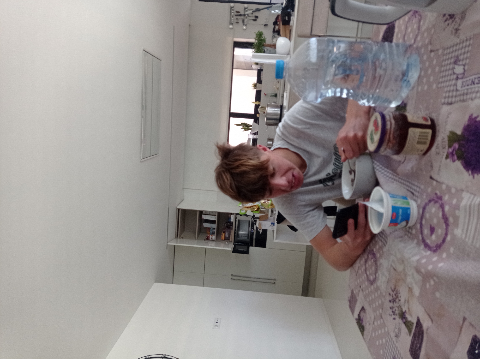The image shows a young person with light brown hair, wearing a grey t-shirt, sitting at a table covered with a patterned tablecloth featuring lavender designs. The individual is holding a black mobile phone in one hand and appearing to prepare food with the other, which involves a jar of jam, a white bowl, and a container of what appears to be yogurt. A large water bottle also rests on the table.
In the background, a kitchen area is visible, featuring white cabinets and appliances, including a microwave and possibly an oven. A window provides natural light to the room, revealing plants and the exterior. Although the location is identified as Felgueiras, Portugal, the image provides no further information to confirm this or provide additional context about the place.