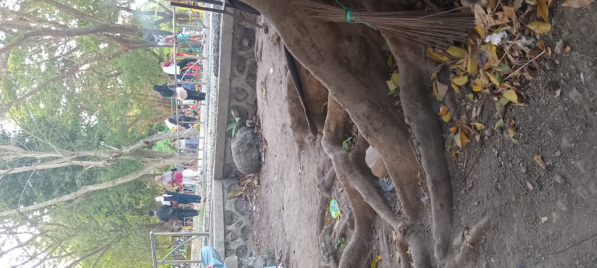 The image depicts a park area with several people engaged in various activities. In the foreground, large tree roots dominate the scene, with a broom made of twigs tied with green string resting against them. The ground is covered in soil, small stones, and fallen leaves, with scattered debris including plastic wrappers and a partially covered object resembling a bowl. A concrete wall with rough-hewn stones separates the foreground from a paved walkway. On this walkway, several individuals are visible: a man and woman in dark clothing, a woman in a pink headscarf and white pants, and others further back, some of whom appear to be seated or standing near playground equipment. The background features lush green trees and foliage, suggesting an outdoor recreational setting. The scene is likely located in Mataram, Indonesia, a city known for its public parks.