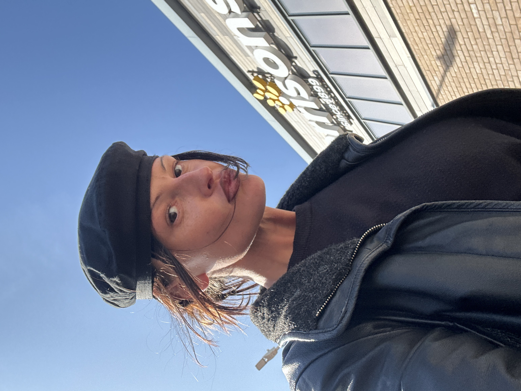 A woman wearing a black beret and black leather jacket poses with her tongue sticking out, likely taking a selfie. She appears to be standing outside a store, as indicated by the sign "Currys" with the tagline "since 1888" visible in the background. The sky is blue. The location is possibly in Folkestone and Hythe District, United Kingdom.