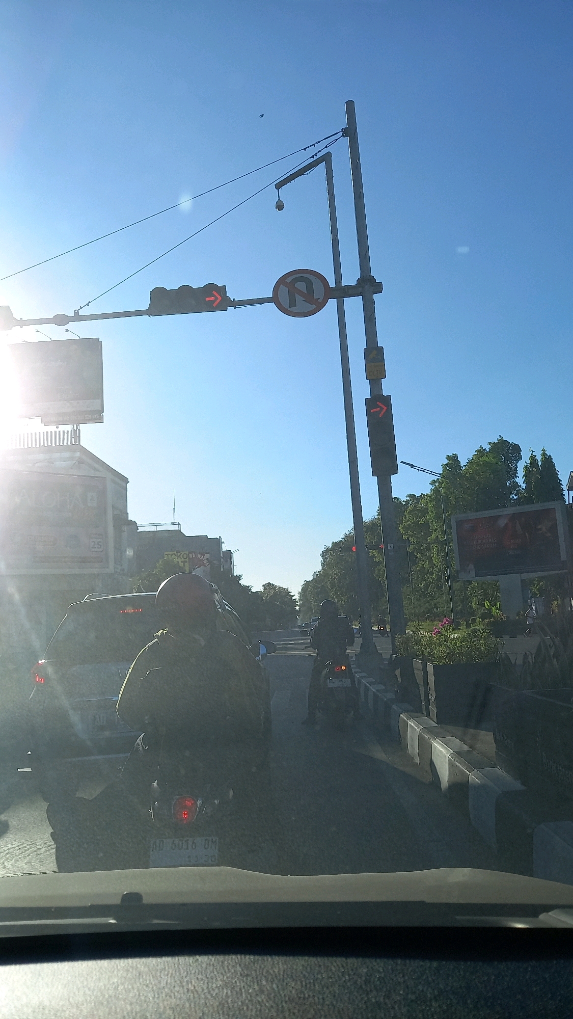 The image captures a traffic scene on a clear, sunny day, likely in the city of Surakarta, Indonesia. The perspective is from inside a vehicle, looking forward. Ahead, there is a traffic light displaying a red arrow, indicating traffic must stop or not proceed in the direction of the arrow. A "no U-turn" sign is also visible. On the right side of the road, a traffic light shows a red arrow pointing to the right. Several motorcycles are in traffic, with one in the foreground in the left lane, and another in the right lane. Buildings, some with signage including "ALOHA" and "TOKO NASIONAL", are visible on the left side of the road. Trees line the right side of the road, with a billboard visible in the distance. The road surface appears to be asphalt. The overall impression is of a busy urban intersection during daytime.