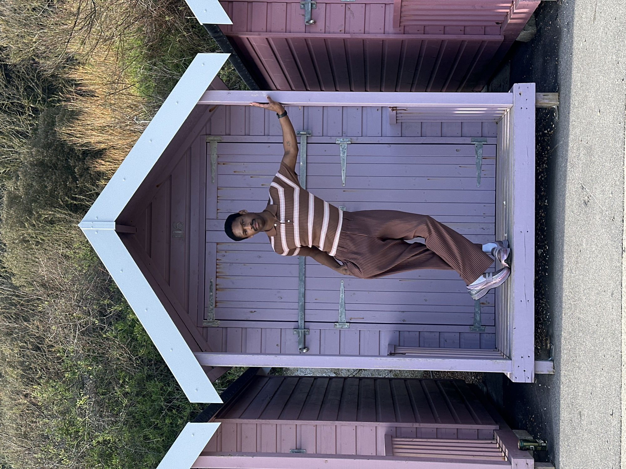 The image captures a slender man posing inside a lilac painted beach hut, one in a row of similar structures. He wears a brown and light pink striped polo shirt and brown pleated pants, accessorized with a black wristwatch and pink sneakers. His left arm is raised, touching the top of the doorframe, while his right hand rests casually on his hip. The huts have white-trimmed gabled roofs. Vegetation borders the huts on one side, and a concrete pathway is visible on the other. The location is most likely Folkestone and Hythe District, United Kingdom, based on the style of the beach huts.