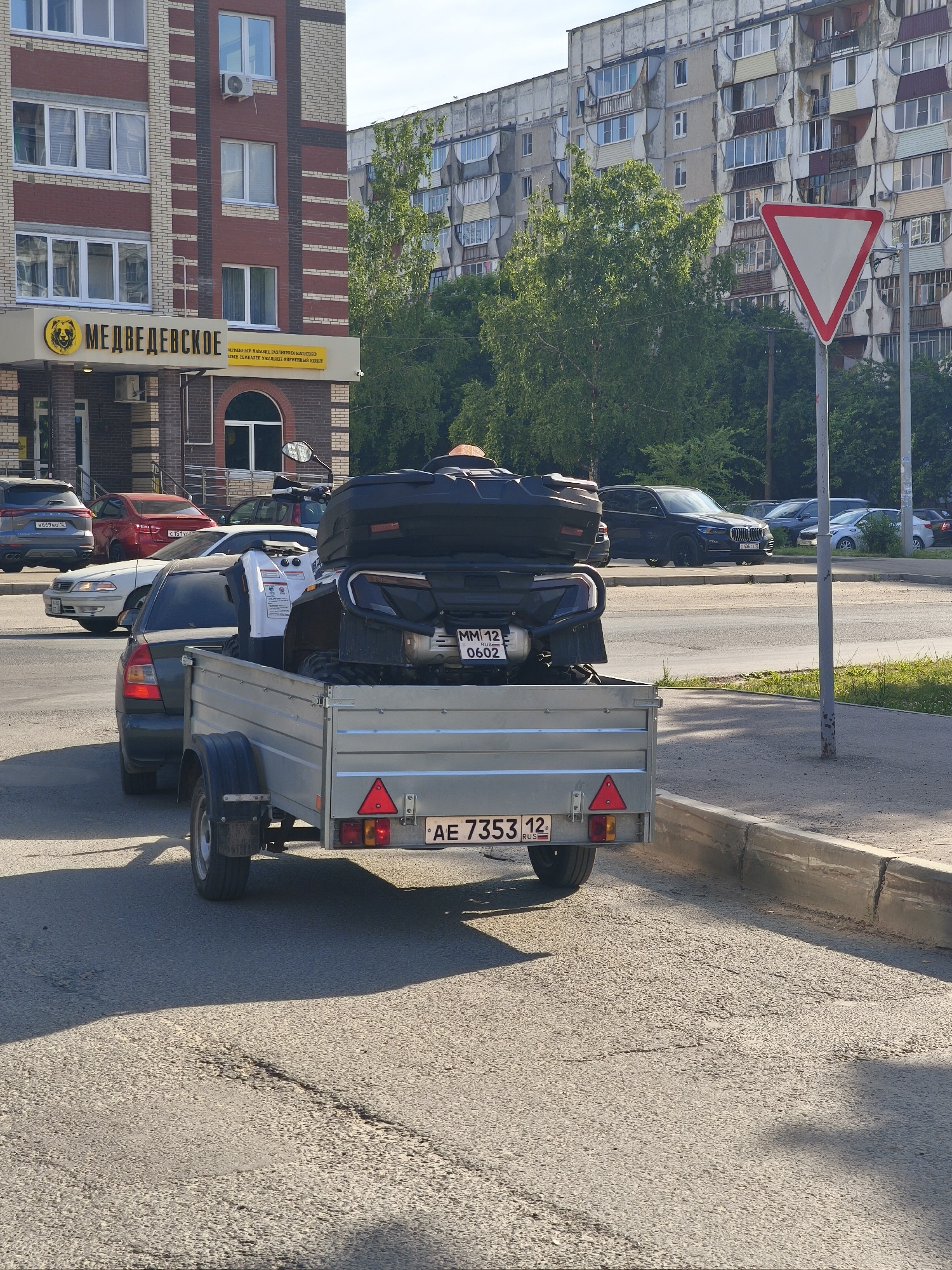 An ATV is being transported on a trailer pulled by a dark colored car. The trailer is silver with red taillights and a license plate reading "AE 7353 12 RUS". The ATV is black with a storage box on the back. The car is a sedan. In the background, there are multi-story apartment buildings and trees. A triangular yield sign is visible on the right side of the image. The scene appears to be on a street in Yoshkar-Ola, Russia, as indicated by the building signage reading "МЕДВЕДЕВСКОЕ" which translates to "Medvedevskoe".