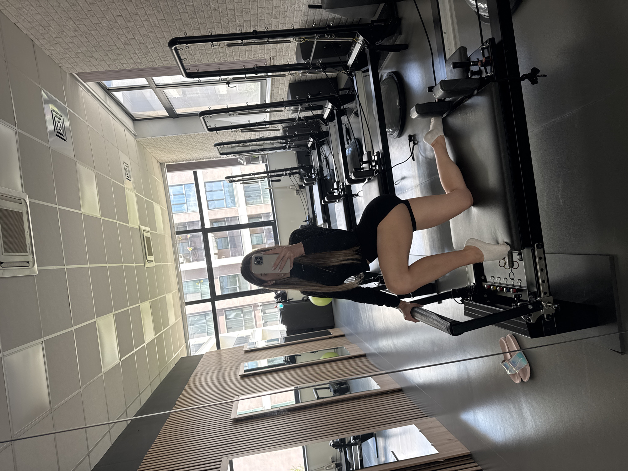 In a Pilates studio in Sofia, Bulgaria, a woman is captured in a selfie while kneeling on a Pilates reformer. She is wearing a black long-sleeved top and shorts, with white socks on her feet. The studio is well-lit with natural light streaming through large windows, offering a view of city buildings. The room is equipped with multiple Pilates reformers, mirrors lining one wall, and a polished concrete floor. A pair of holographic slides are visible on the floor to the left of the reformer.