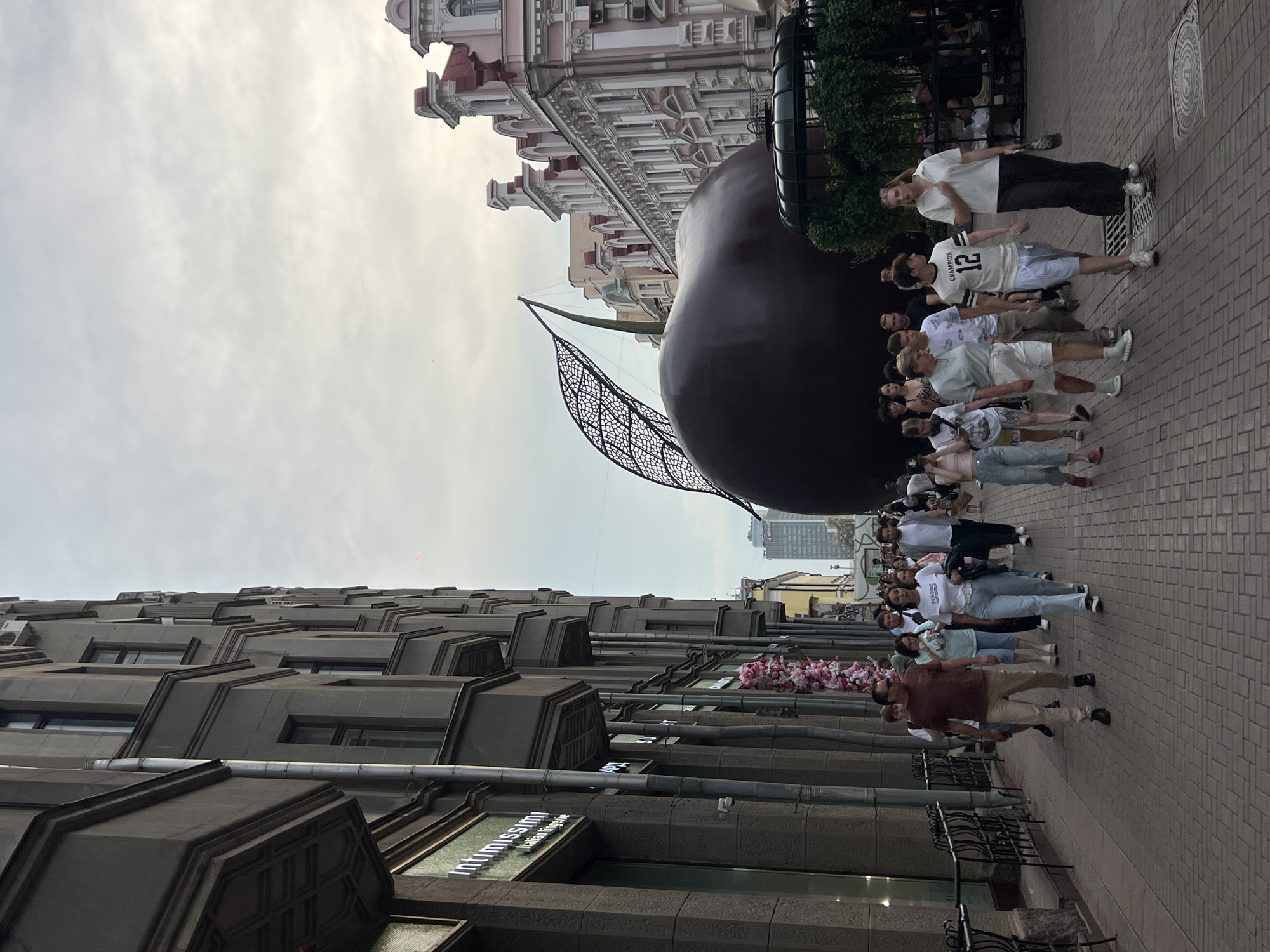 The image depicts a street scene with a large, dark red, sculptural cherry and a leaf. A group of people are walking on a brick-paved sidewalk alongside buildings. The architecture of the buildings varies, with some appearing older with ornate details and others being more modern and taller in the background. The sky is overcast. The street appears to be a pedestrian walkway with shops lining one side. The scene suggests a daytime urban environment. One of the buildings has signage for "Intimissimi Italian Lingerie." A woman in the foreground is wearing a white t-shirt with "SEASIDE" printed on it and light blue jeans, and a man next to her is wearing a maroon t-shirt and khaki pants.