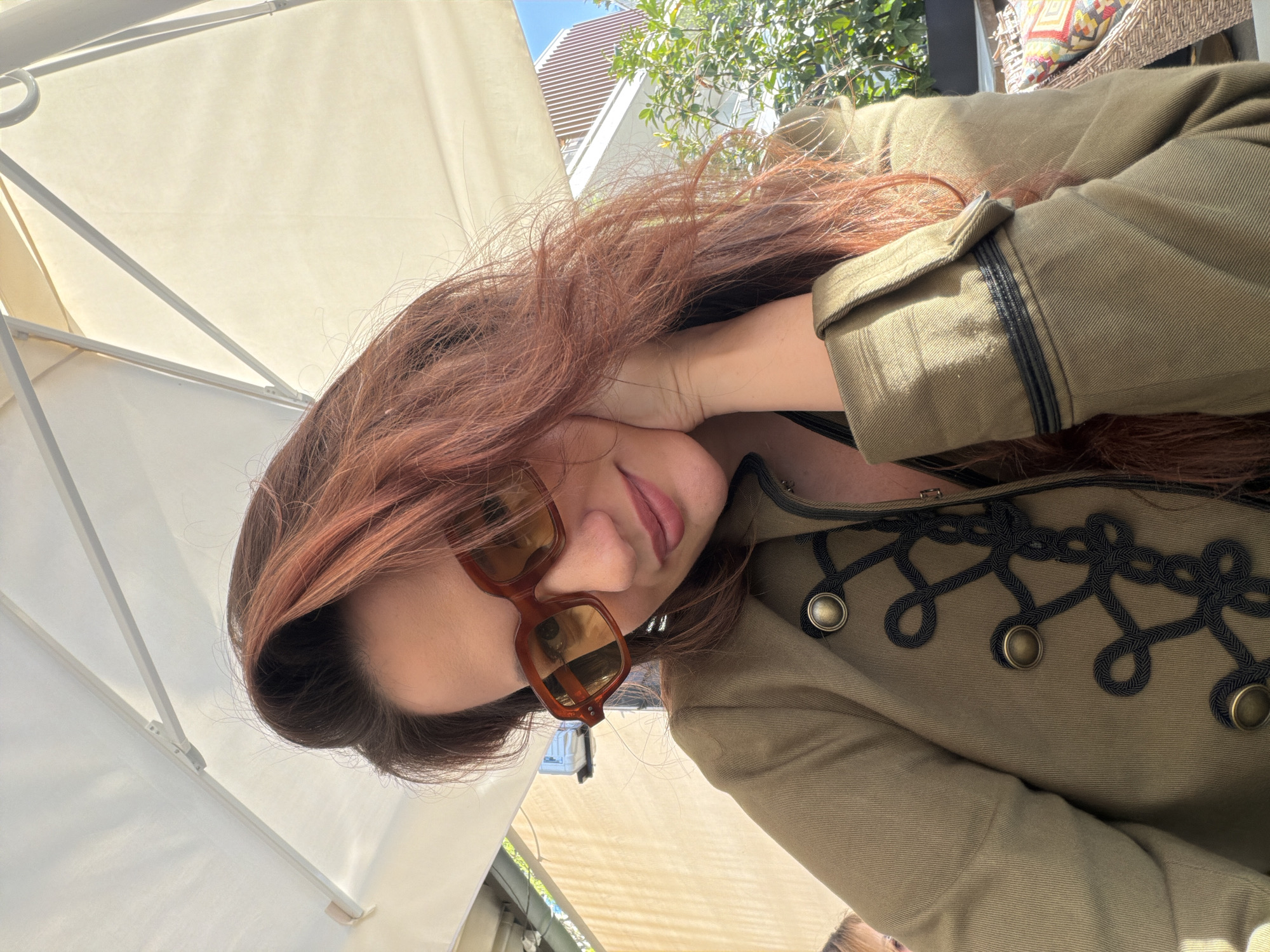 A woman with long, flowing reddish-brown hair is captured in a close-up, outdoor portrait in Belgrade, Serbia. She looks towards the camera with a soft smile, her head tilted, and one hand gently resting near her neck. She wears stylish brown-framed sunglasses with tinted lenses. Her attire consists of an olive-green jacket, distinctively adorned with an intricate black cord pattern across the chest, featuring multiple loops and several round, bronze-toned buttons. The jacket also has folded cuffs with black stitching.
The setting is a sunlit outdoor area, likely a cafe or restaurant patio. A large white umbrella or parasol provides shade in the background. Beyond her, elements of an urban environment are visible, including green foliage from trees or bushes and a modern building with slatted architectural details under a clear sky. A wicker chair with a patterned, colorful cushion is partially visible in the background. The bright daylight suggests a sunny day. No visible text is present in the image.