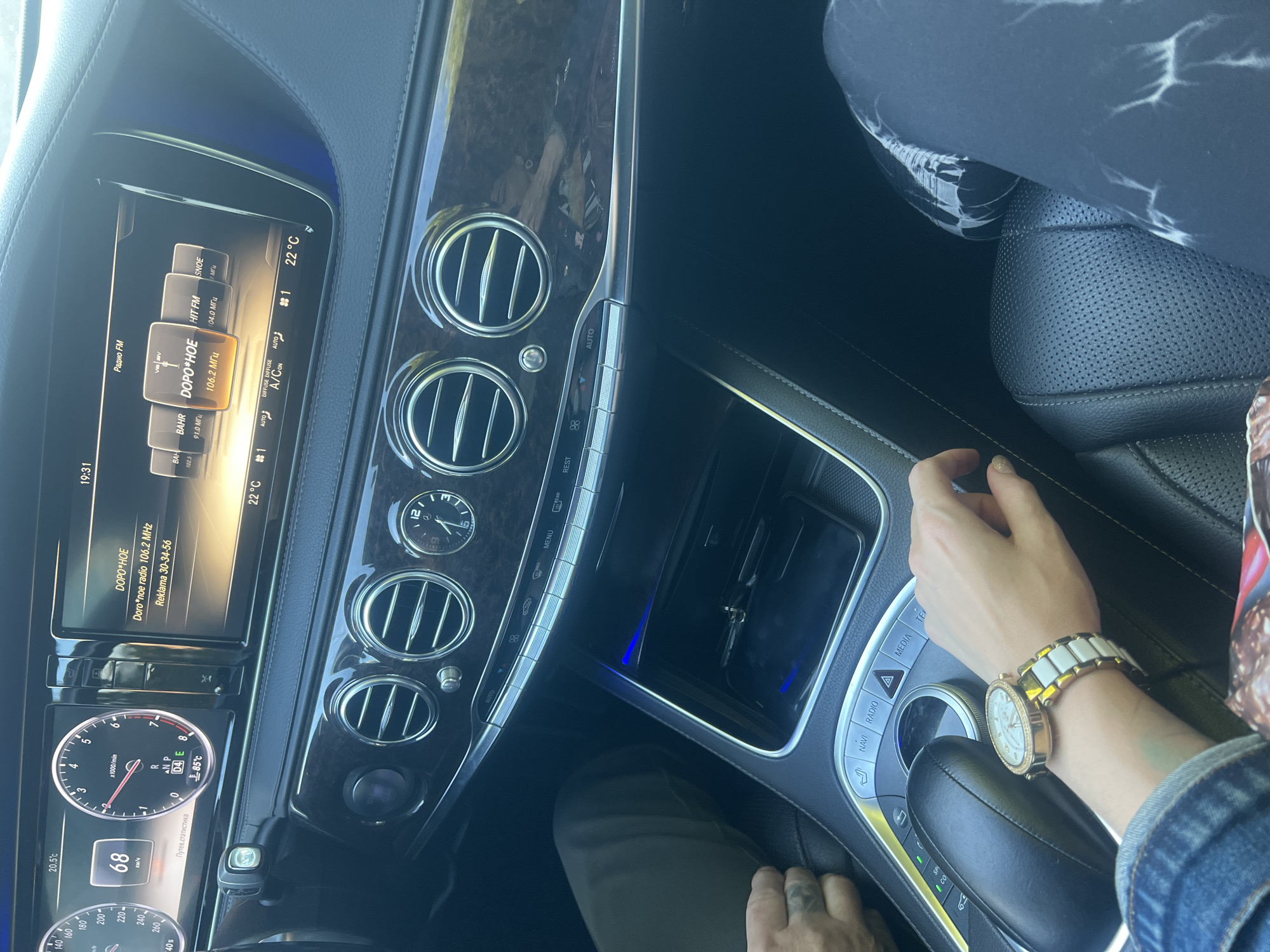 The image shows the interior of a Mercedes-Benz S-Class, a luxury sedan, with a focus on the dashboard and center console. A woman's arm, adorned with a prominent gold and white watch, rests on the center console. Her hand is lightly touching the console. Next to her, a man's hand with a visible tattoo is resting on the armrest. The car's digital display shows the time as 19:31 and the temperature as 20.5°C outside. The infotainment screen is tuned to "DORO*HOE" radio on 106.2 MHz. The center console features climate control buttons, ventilation grilles, and a gear selector. A smartphone and keys are visible in the storage compartment below the climate controls. The ambient lighting in the car is a soft blue. The image appears to have been taken while the car is stationary, likely parked. The backdrop of Krasnoe-na-Volge, Russia, suggests a local setting, possibly a residential area or a public space within the town.