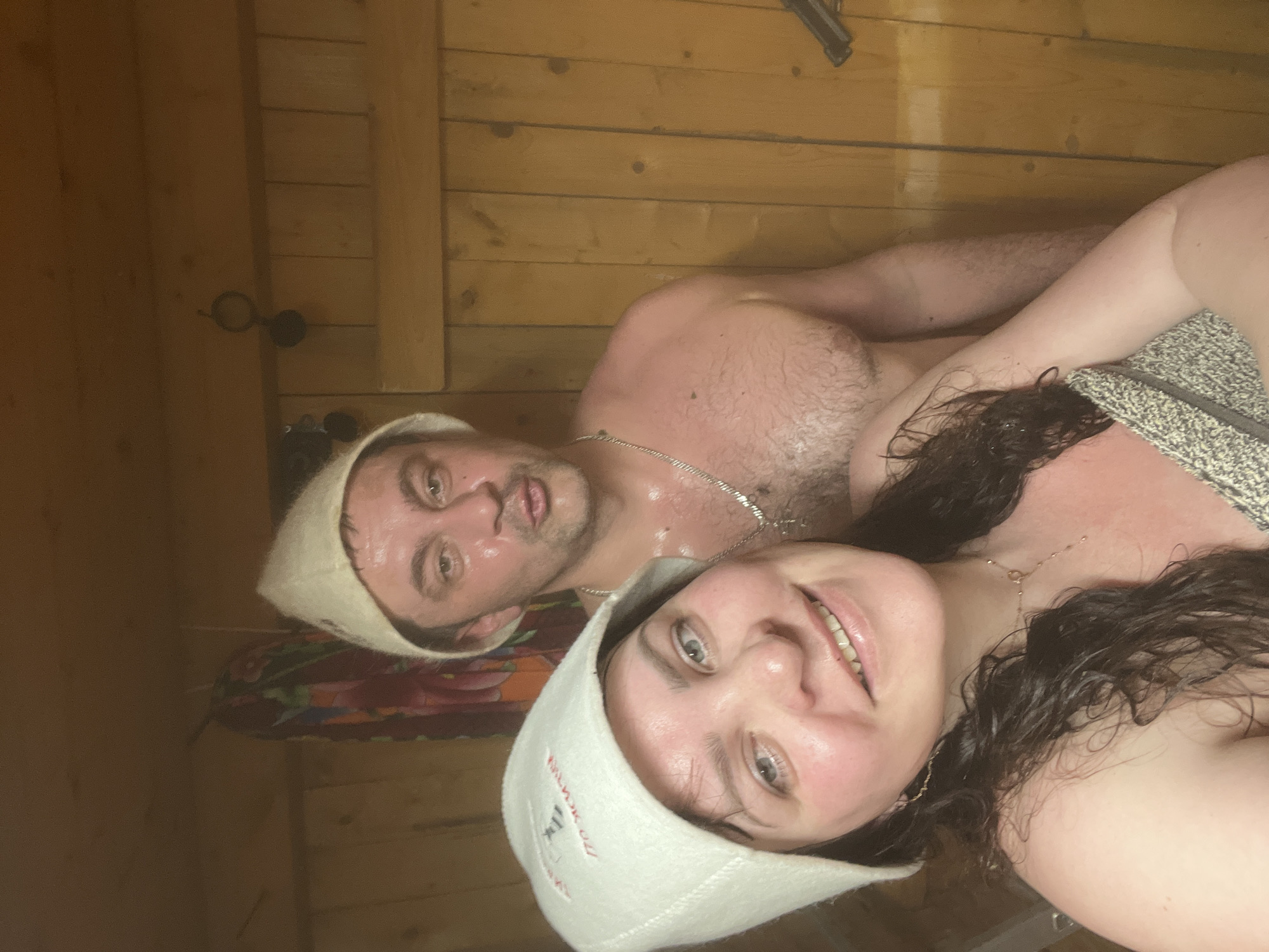 The image displays a close-up selfie of a man and a woman inside what appears to be a sauna or banya. Both individuals are looking directly at the camera, with the woman positioned in the foreground and the man behind her to the left.
The woman has dark, wet hair and a clear, joyful smile on her flushed, sweaty face. She wears a white felt sauna hat with red Cyrillic text and a small graphic. The visible text on her hat reads "БАНЯ" (Banya/Sauna) and "С ЛЁГКИМ ПАРОМ" (S Lyogkim Parom), a common Russian wish for a good steam. She also wears a delicate gold-colored necklace and her skin is visibly moist from the heat. A light-colored, patterned towel is draped over her left shoulder and chest.
The man, partially visible behind the woman, also has a flushed and sweaty complexion. He is wearing a plain white felt sauna hat and a silver chain necklace. His bare torso shows chest hair. His expression is content, with a slight smile.
The setting is characterized by light-colored wooden planks forming the walls, indicative of a sauna. A faint atmospheric haze suggests steam or heat. Behind the man's head, a colorful patterned fabric, featuring shades of red, green, and orange, is visible. To the far left, a black door handle with a circular grip is affixed to the wooden wall. The overall scene conveys an atmosphere of relaxation and enjoyment in a hot, steamy environment.