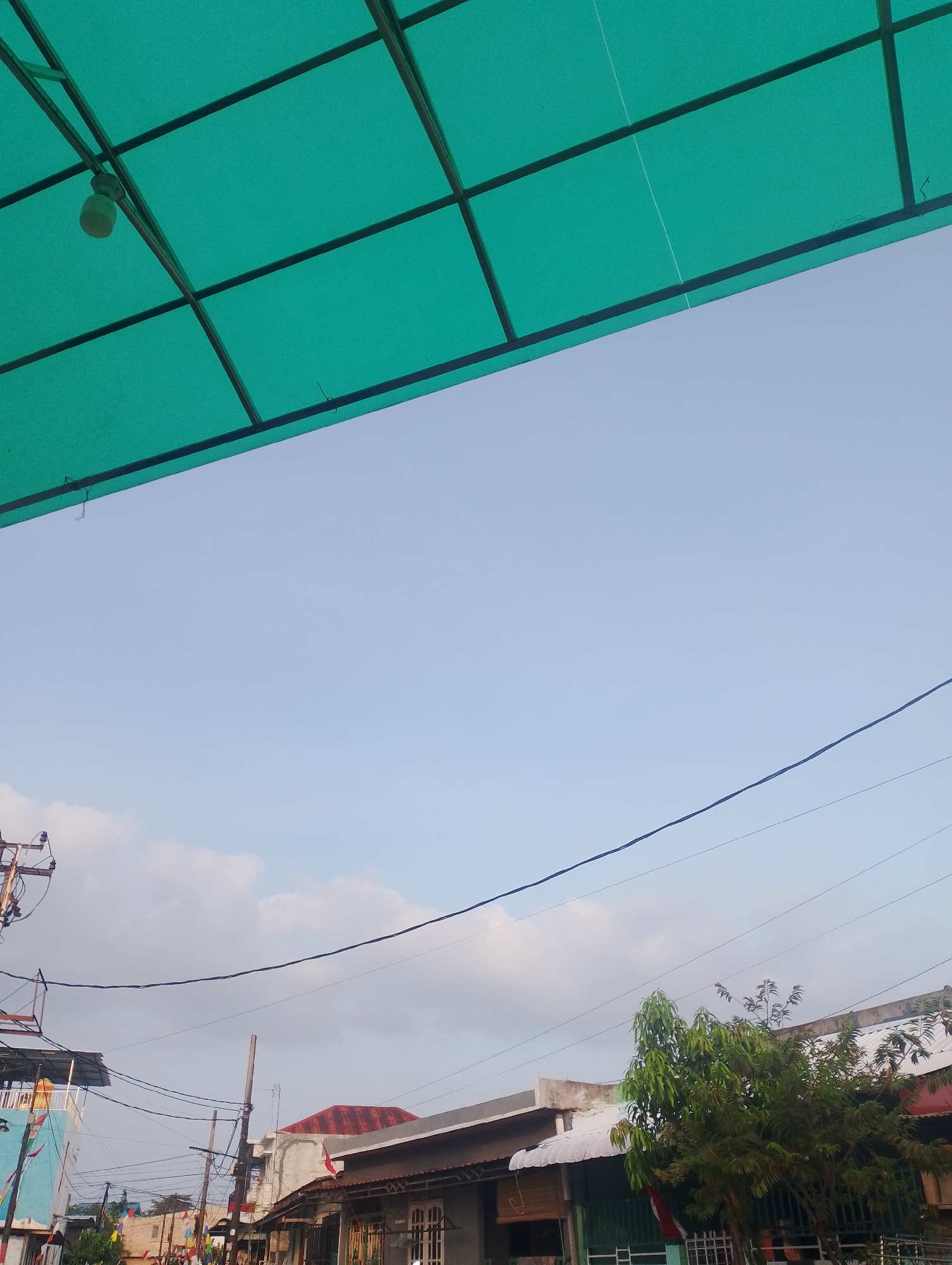 The image captures a street view in Batam, Indonesia, under a pale blue sky with scattered clouds. In the foreground, a green translucent roof supported by dark metal bars is visible at the top, with a light bulb hanging from it. The middle ground shows a network of electrical wires crisscrossing overhead, supported by wooden and metal poles. Below the wires, residential buildings with various roofing materials, including corrugated metal and red tiles, line the street. One building features a blue and white facade with colorful pennants strung along its side, suggesting a local celebration or event. A leafy green tree stands in front of some of the buildings. The scene appears to be a typical residential or commercial street in a tropical climate, likely during daylight hours. There are no people visible in the image.