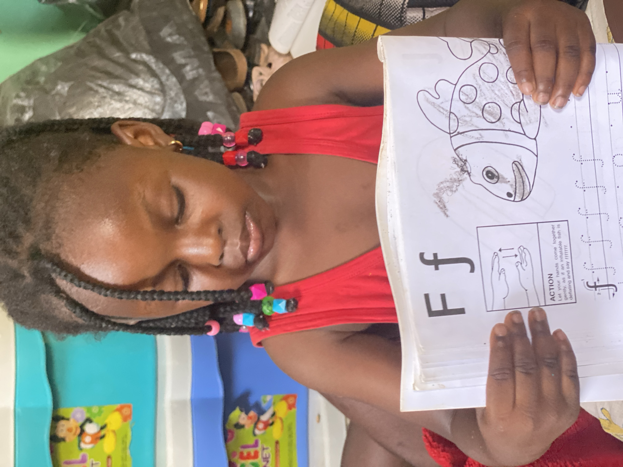 A child with braids and colored beads is lying on their side, holding a book open to a page with the letter "F" and a drawing of a fish. The child is wearing a red shirt. The background includes furniture with cartoon character designs and what appears to be bedding and shoes. While there is no specific location information, the OCR mentions Rido, Nigeria. However, no information in the image allows that location to be confirmed.